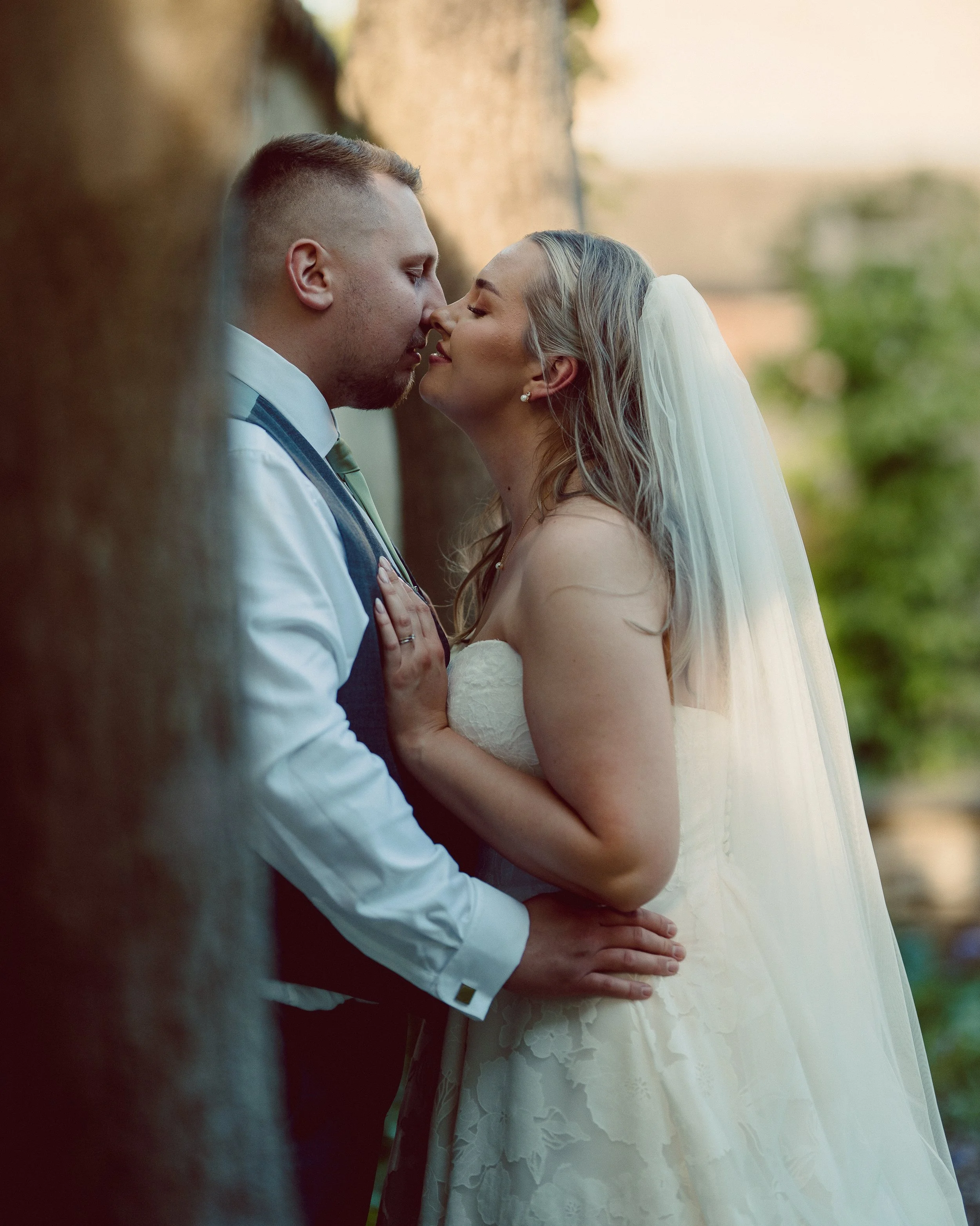 A bride and groom share an intimate moment, about to kiss, outdoors during their wedding. The groom has a short haircut and a beard, wearing a white shirt and a vest. The bride has wavy gray hair, wearing a strapless lace wedding dress and a veil. Th