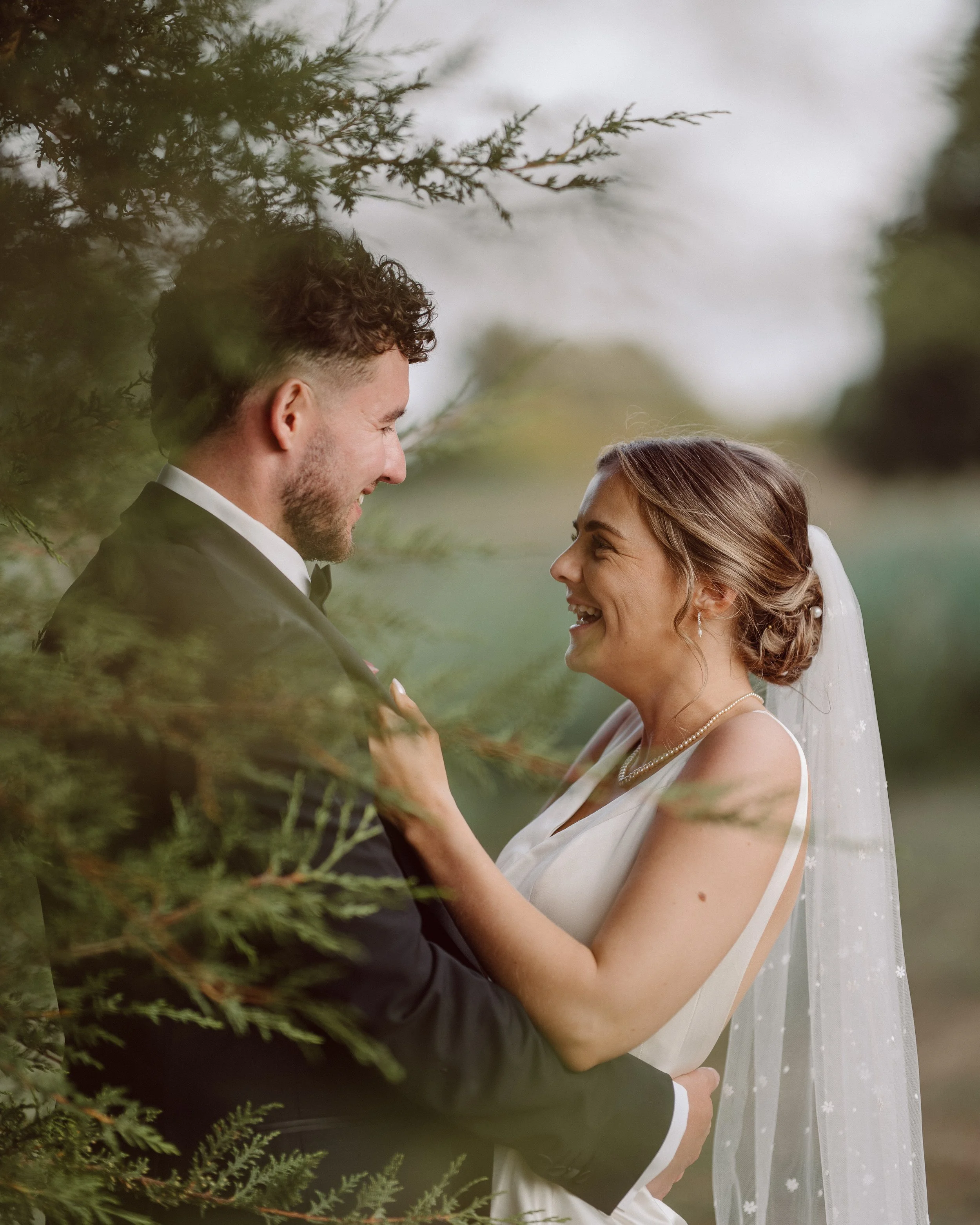 A couple is smiling and looking at each other outdoors, surrounded by trees and greenery, dressed in wedding attire. The man is wearing a black tuxedo and the woman is in a white wedding dress with a veil.