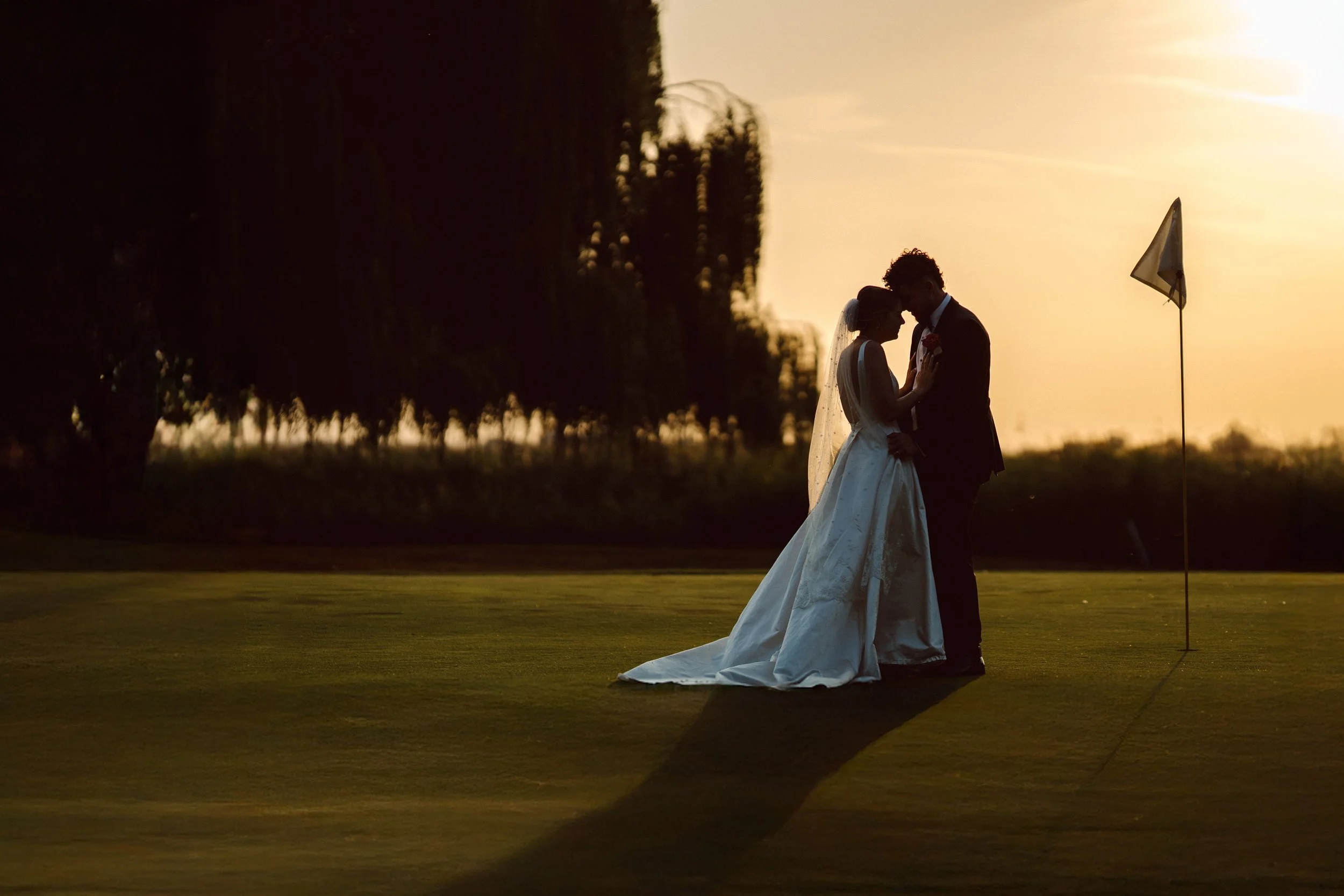 A bride and groom are standing close together outdoors during sunset, with their foreheads touching, in a romantic moment. The bride is wearing a white wedding gown with a long train and veil, and the groom is in a dark suit. There is a flag nearby, 