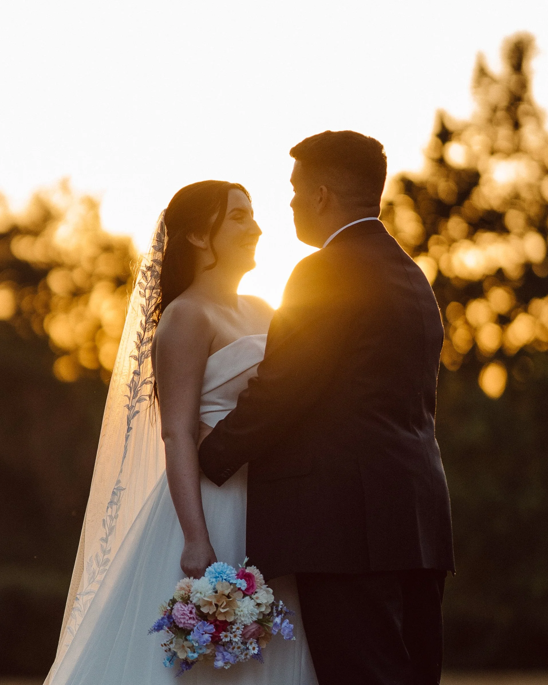A bride and groom facing each other outdoors during sunset, with the bride holding a colorful bouquet of flowers. The background shows blurred trees and the warm glow of the setting sun.