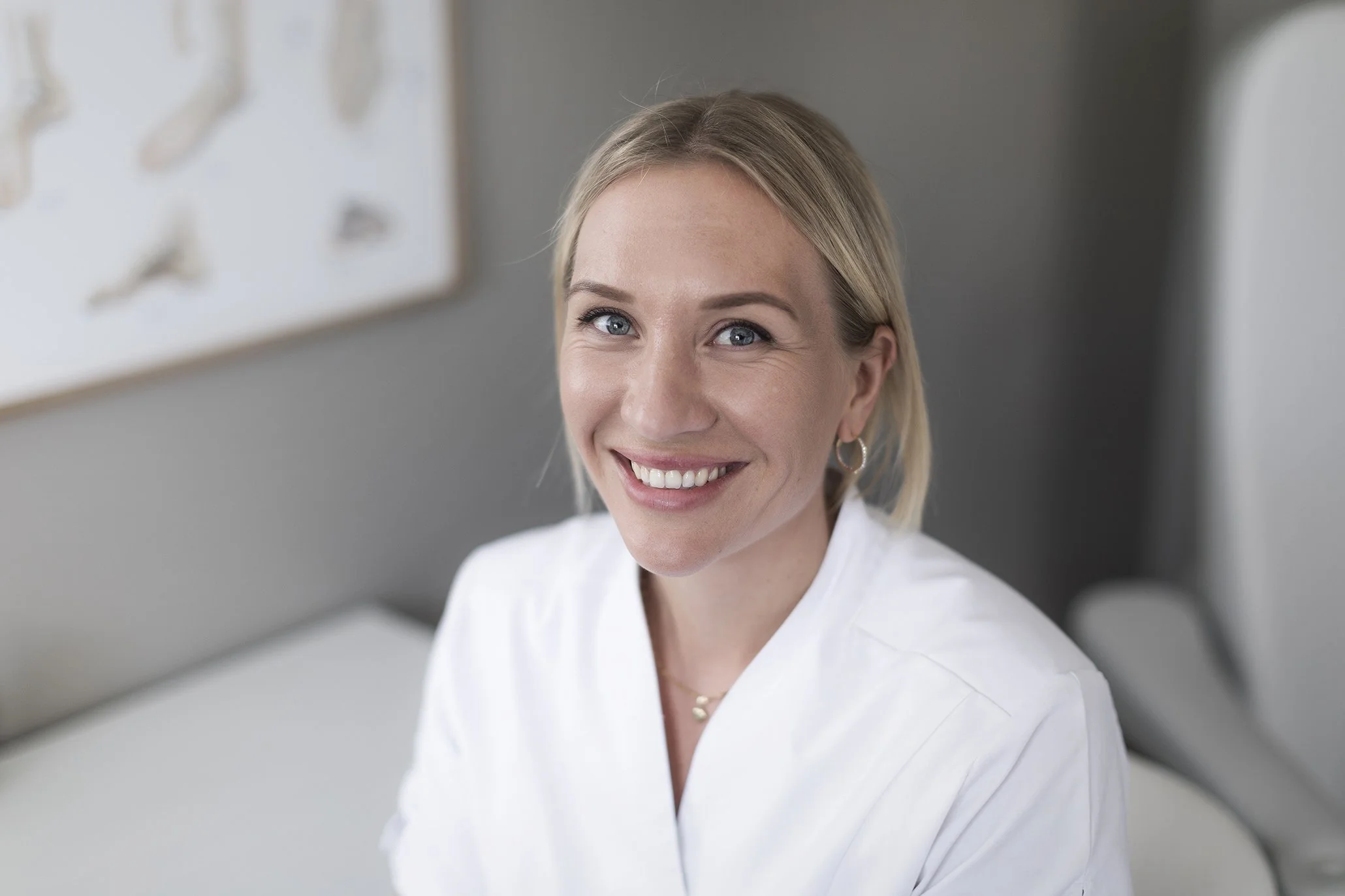 En kvinne med blond hår og hvitt smilende ansikt, kledd i hvit sykehusinstitusjonsfrakk, sitter i et rom med grå vegg og bilder eller ornamenter på veggen.