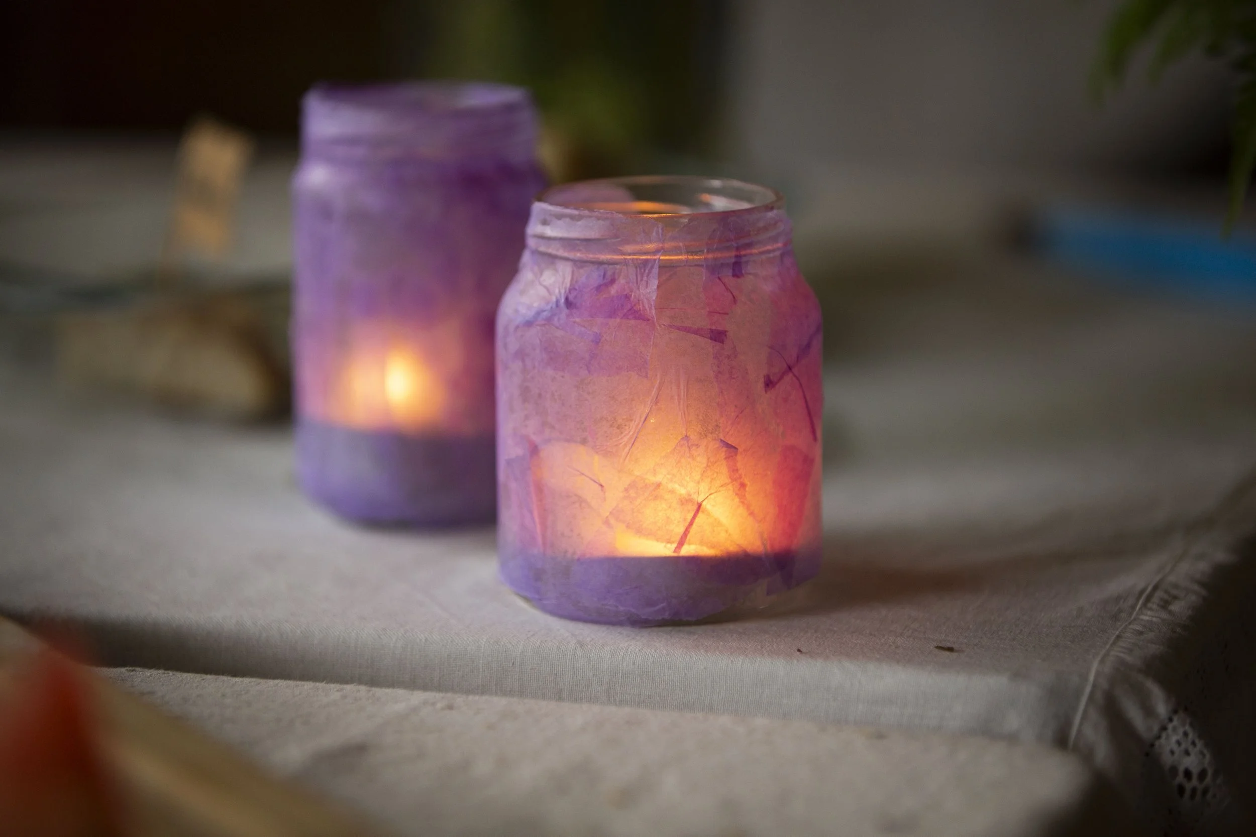 Zwei Kerzen in dekorativen Gläsern mit lila und pinken Tönen auf einem Tisch, die warmes Licht ausstrahlen.