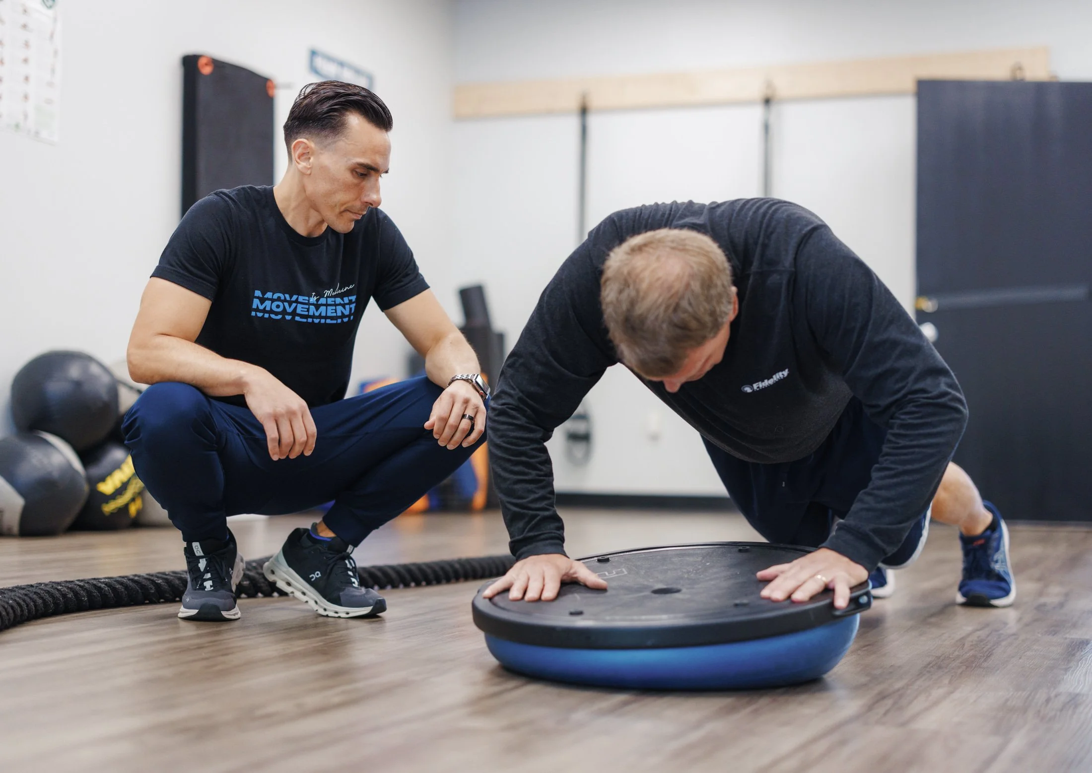 A man performing a push-up on a balance board while a trainer crouches nearby in a gym setting.