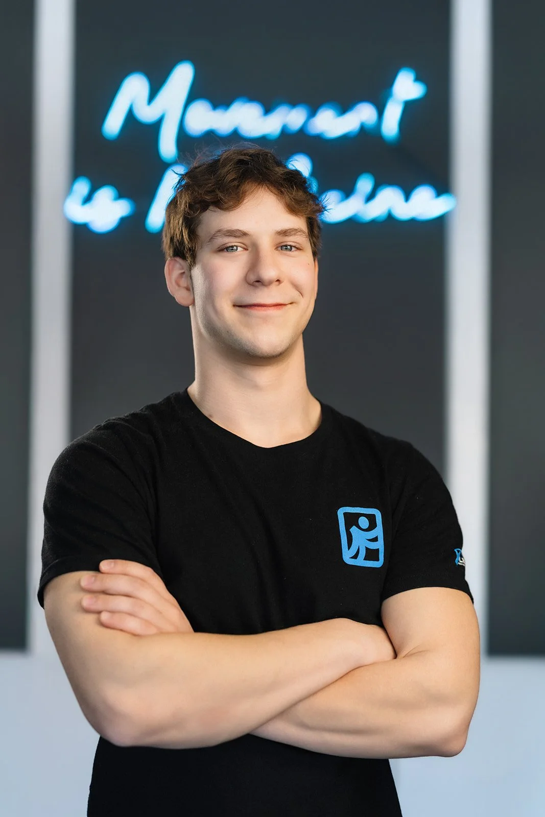 A young man with glasses and dark hair, smiling, standing in front of a neon sign that reads, 'Movement is Medicine,' inside a modern interior with gray walls, a fan, and a small table with brochures.