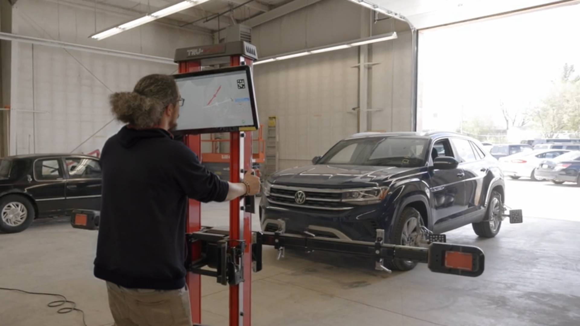 A person is operating a recalibration on a black Volkswagen SUV inside a testing facility, with a large open garage door in the background.