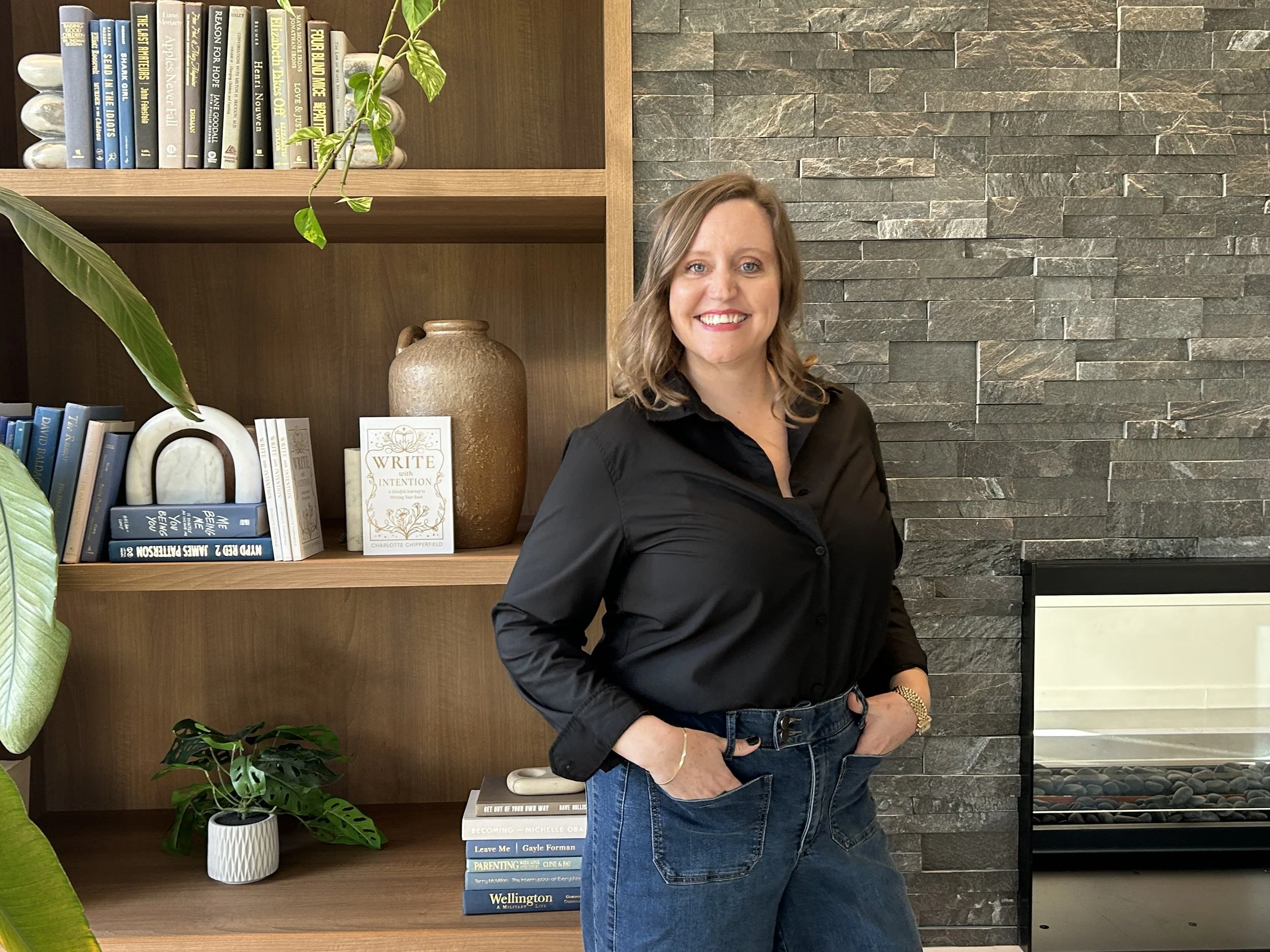 A woman with shoulder-length brown hair, wearing a black blouse and blue jeans, standing indoors in front of a bookshelf with various books, a plant, and decorative items. She is smiling and looking at the camera.