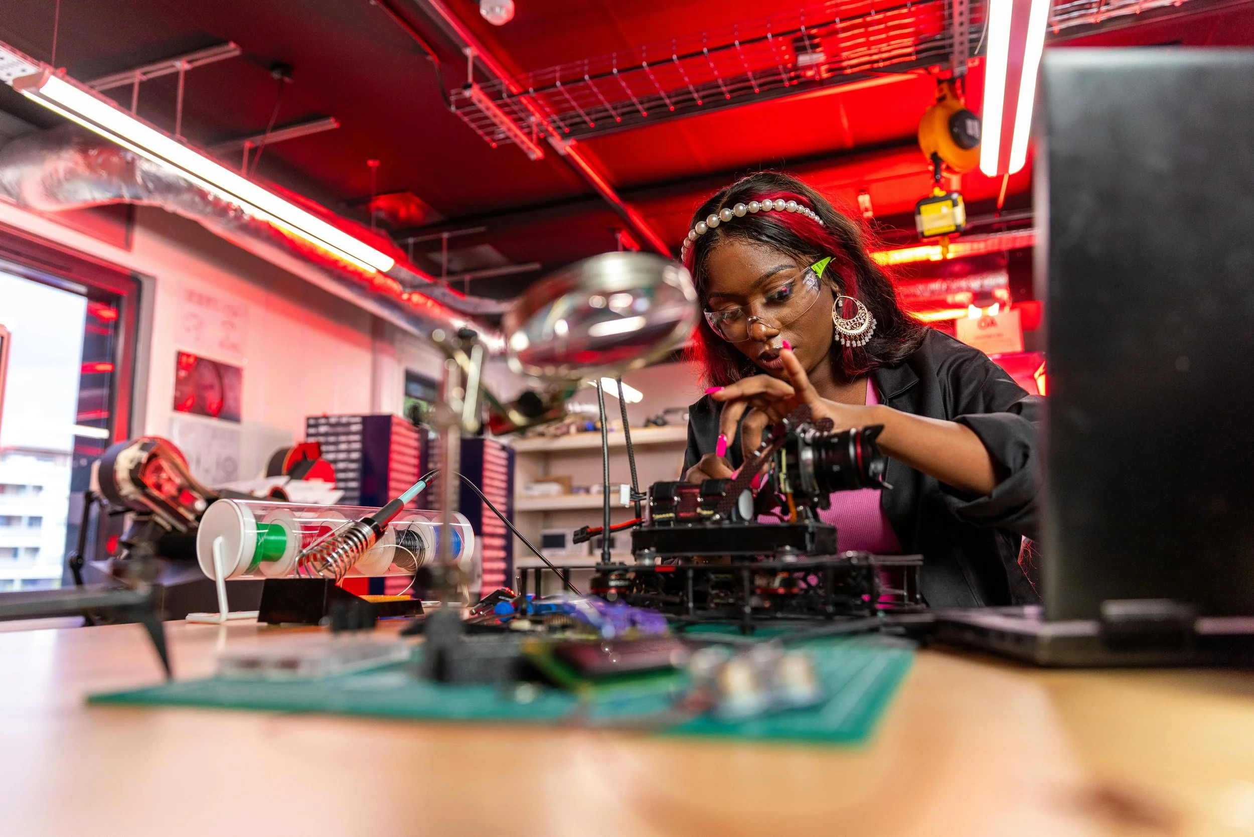 Young woman working on a drone in a workshop with red lighting, safety goggles, and jewelry.