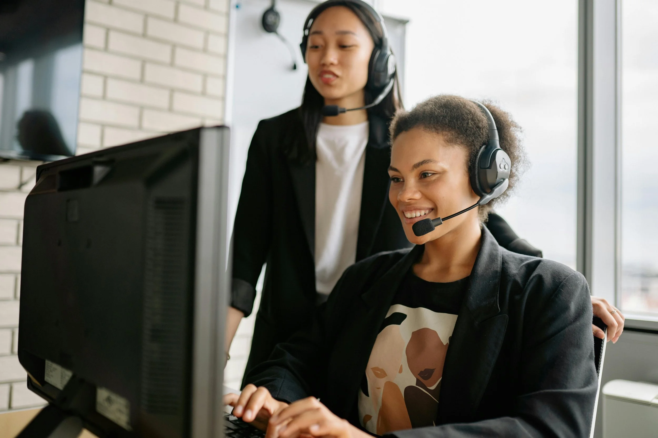 Two women wearing headsets at a computer, working together in an office with large windows and a brick wall in the background.