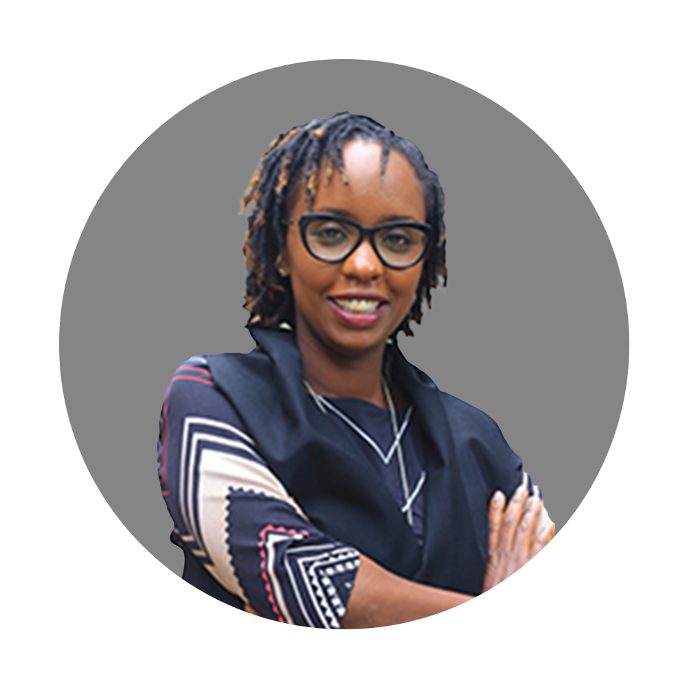 A woman with glasses and dreadlocks smiling with arms crossed, wearing a patterned shirt and silver necklace.