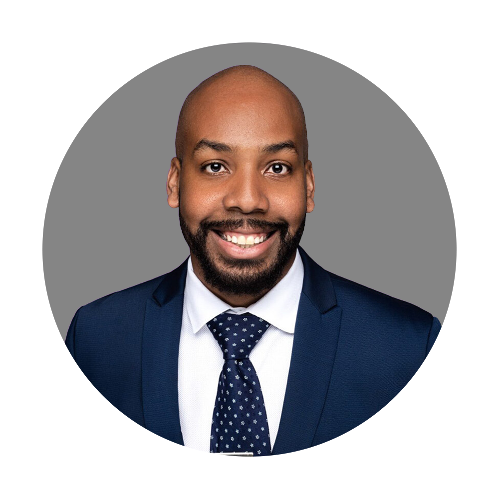 Headshot of a man with a bald head, beard, and mustache, dressed in a blue suit, white shirt, and patterned tie, smiling against a gray and black background.