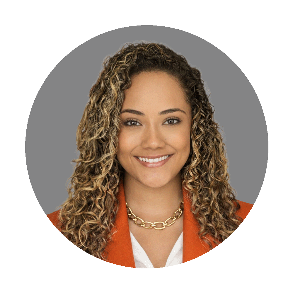 A woman with curly blonde hair smiling in a professional headshot, wearing a gold chain necklace and an orange blazer against a gray background.