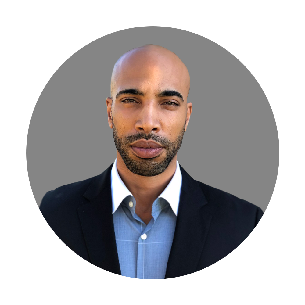 Close-up portrait of a bald man with a beard wearing a dark suit jacket and a light blue dress shirt against a gray background.