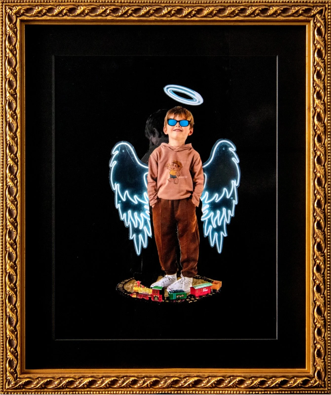 A young boy wearing sunglasses and a hoodie stands in front of a neon light display of angel wings, a halo, and clouds, with toy trains on the ground beneath him, all framed by a gold ornate picture frame. collage by Fabio Borges - Cura Series