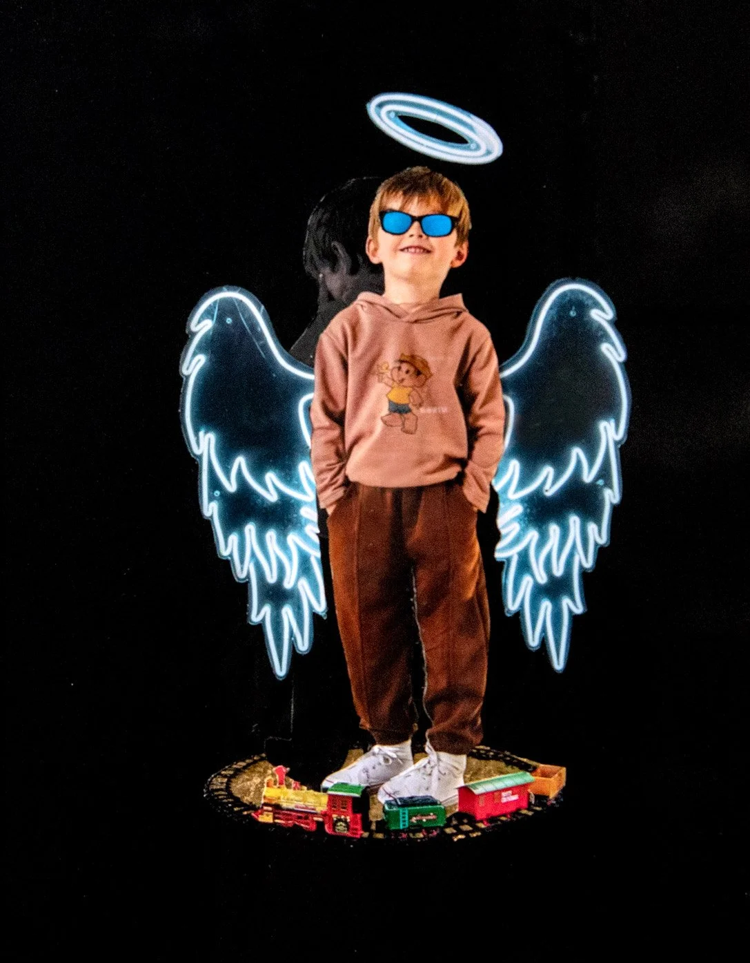 A boy wearing sunglasses and a hoodie with a cartoon character standing in front of a neon sign of angel wings and a halo, with toy train tracks and trains at his feet. collage by Fabio Borges - Cura Series