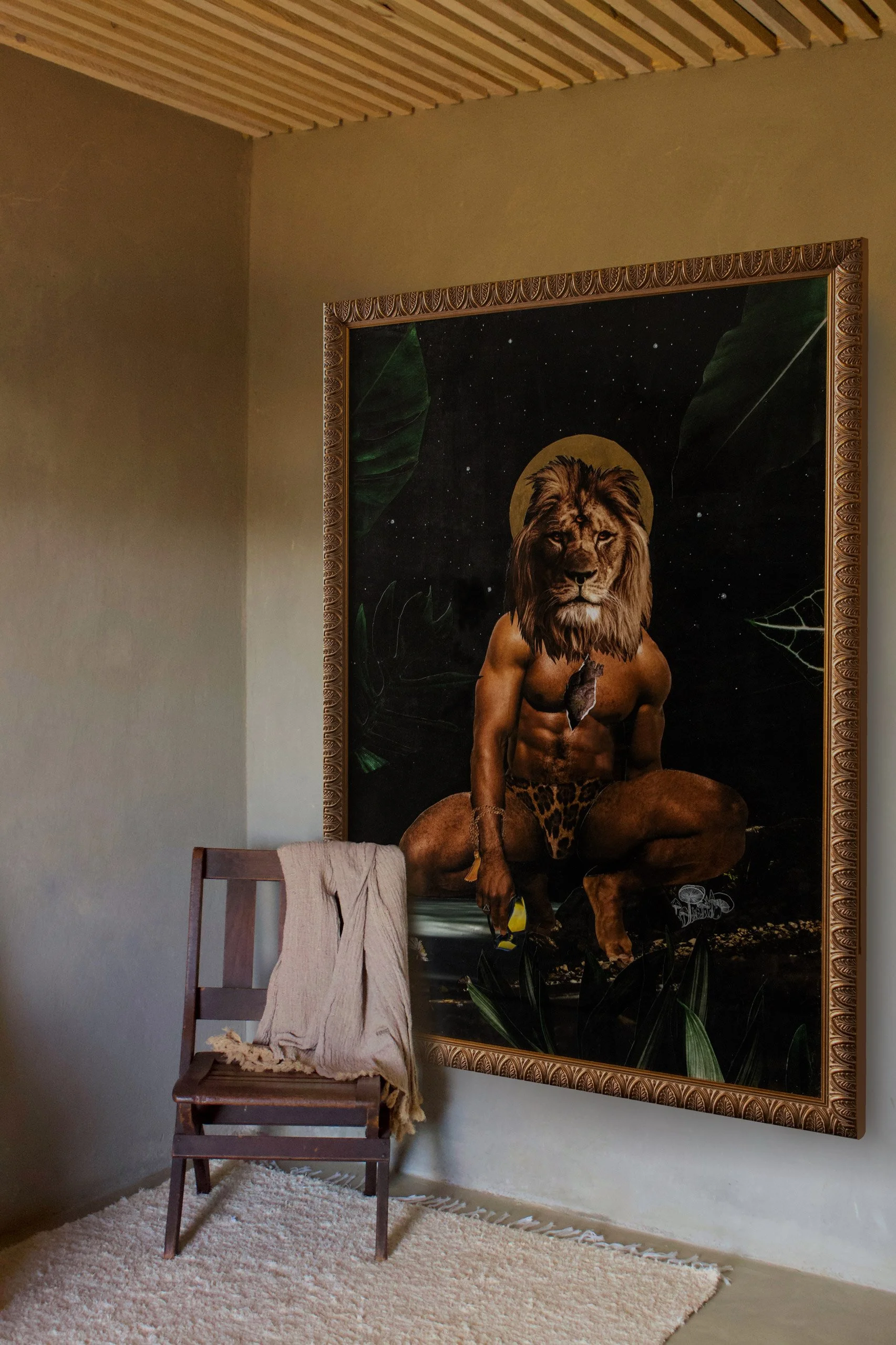 A room with a wooden chair draped with a beige cloth and a large, framed artwork featuring a humanoid lion figure with a human body, a lion's head, and dressed in leopard print shorts, set against a dark, starry background with large green leaves.
