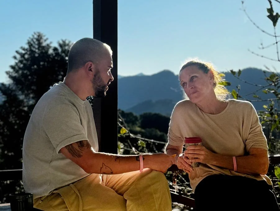 A young man and an older woman sit outdoors, holding hands, with mountains in the background during daytime.