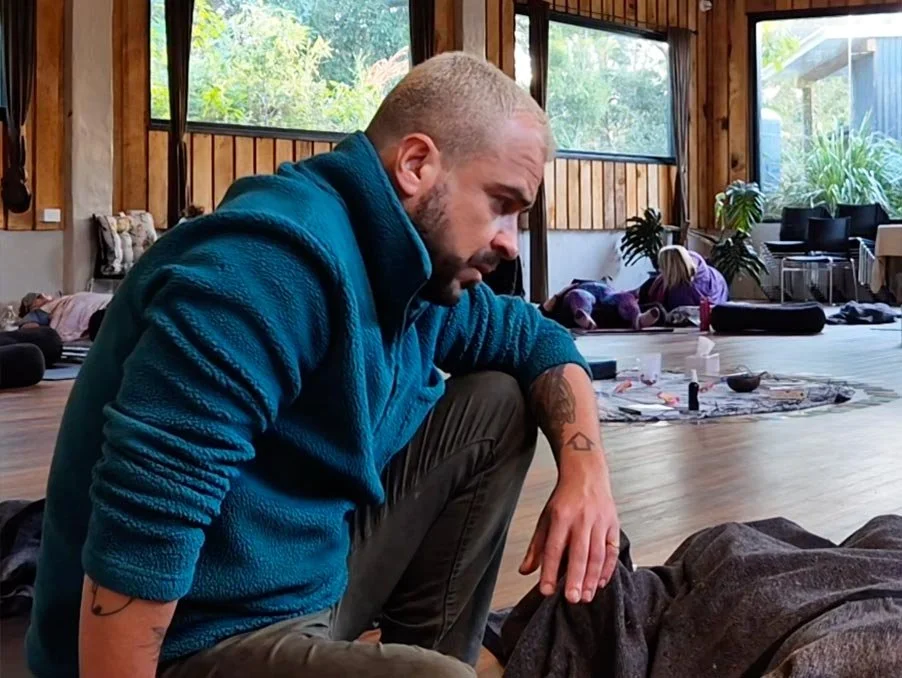 A man with a buzz cut and beard, wearing a blue fleece jacket, is kneeling and looking down at a person lying on the floor. The setting appears to be a yoga or meditation class in a bright, wooden-walled room with large windows showing greenery outside.