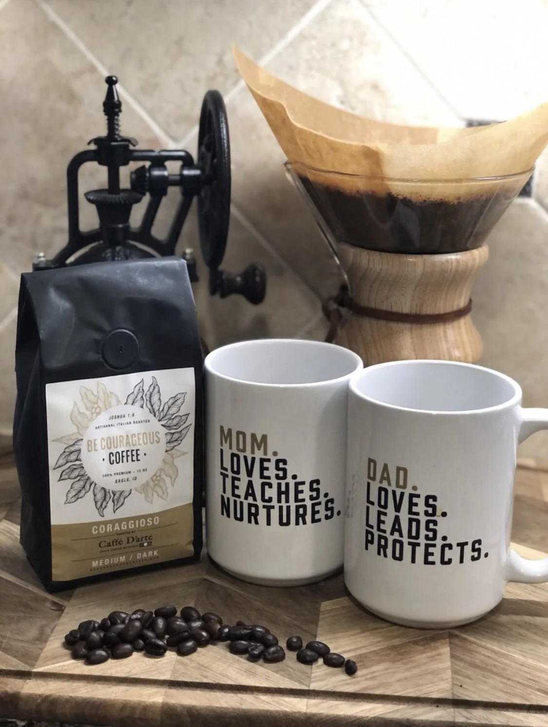 Coffee setup with a bag of Be Courageous Coffee, coffee mill, Chemex with filter, two mugs with text, and coffee beans on wooden surface.
