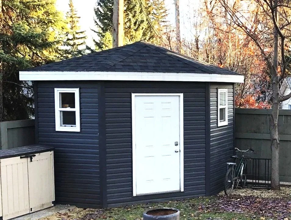 A custom-built, octagonal shed with a white door and two windows, situated in a backyard with a bike leaning against the fence and trees with autumn leaves.