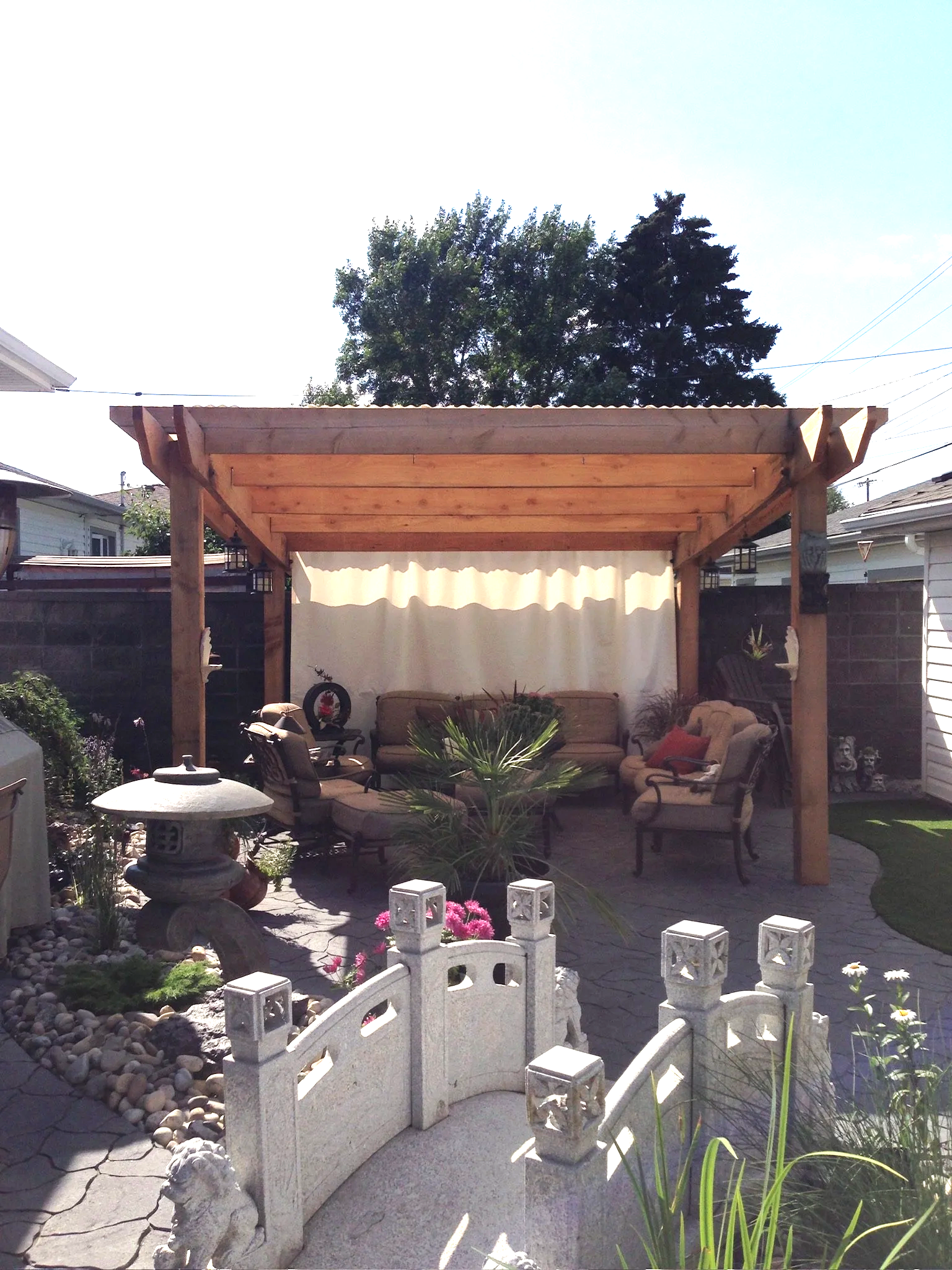 A backyard patio with a custom wooden pergola, outdoor seating, a small white decorative bridge, a rock garden, potted plants, and a large tree in the background.