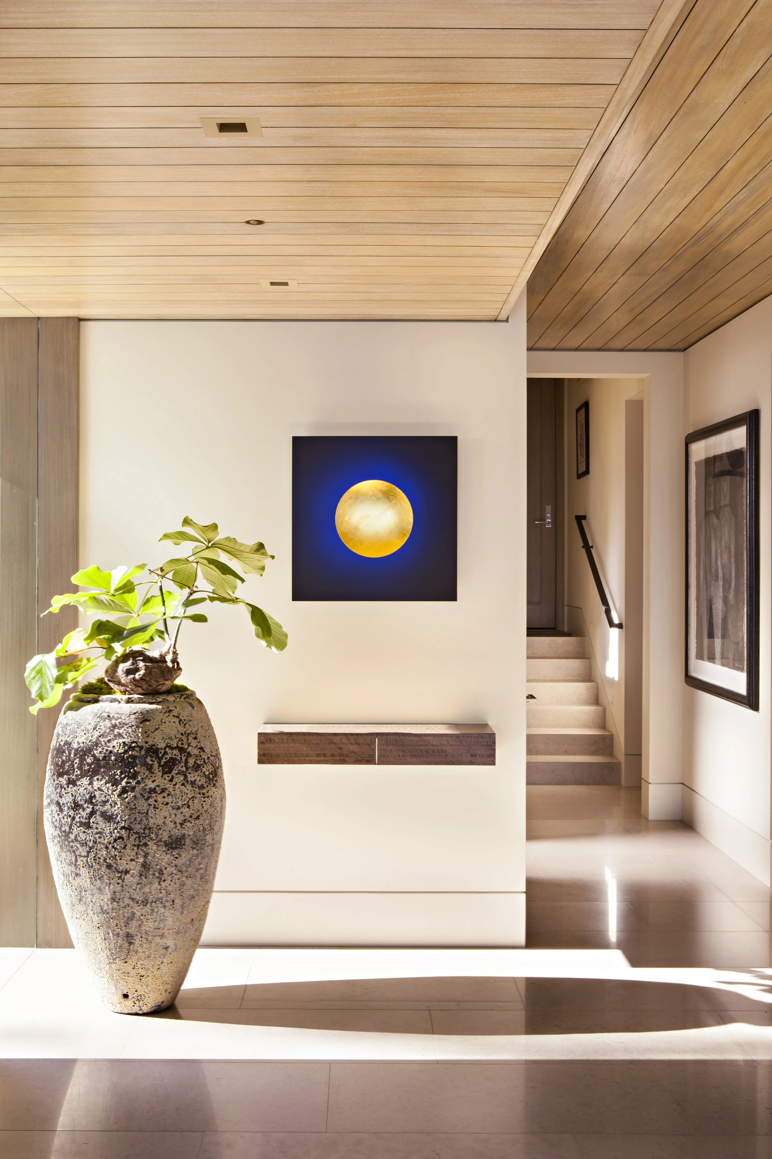 Modern interior space featuring a tall, weathered ceramic planter with green leafy plant, a wall-mounted abstract art piece with a moon-like circle, and a staircase leading to an upper level, with wooden ceiling panels and framed artwork on the wall.