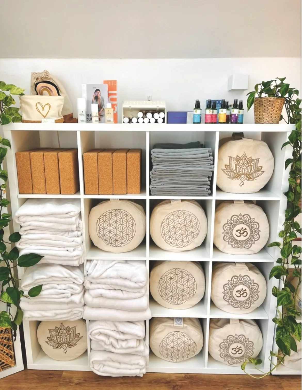 Regal mit Yoga- und Meditationsartikeln, darunter gefaltete Decken, gerollte Kissen mit Chakra-Designs, ätherische Öle, Pflanzen und kleine Verpackungen.