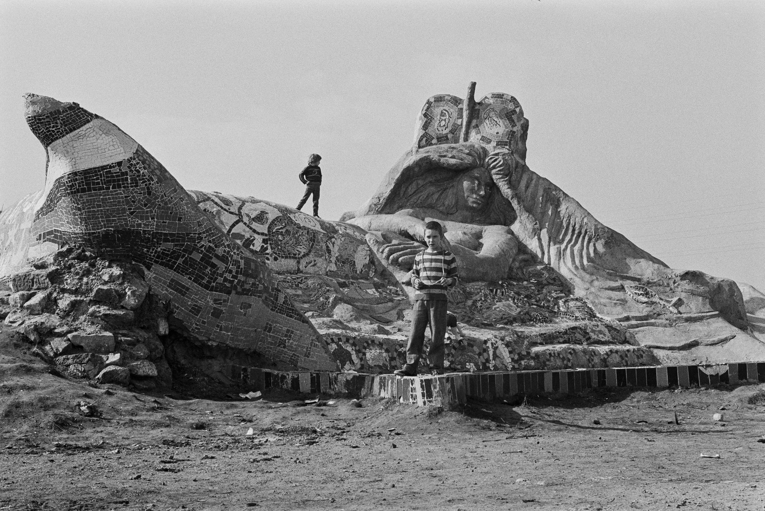 Image description: black and white landscape photograph of the Bingham Mermaid sculpture. A child with light skin, very short dark hair, and dressed in a stripy light and dark top and grey trousers stands at the foot of a large concrete sculpture of