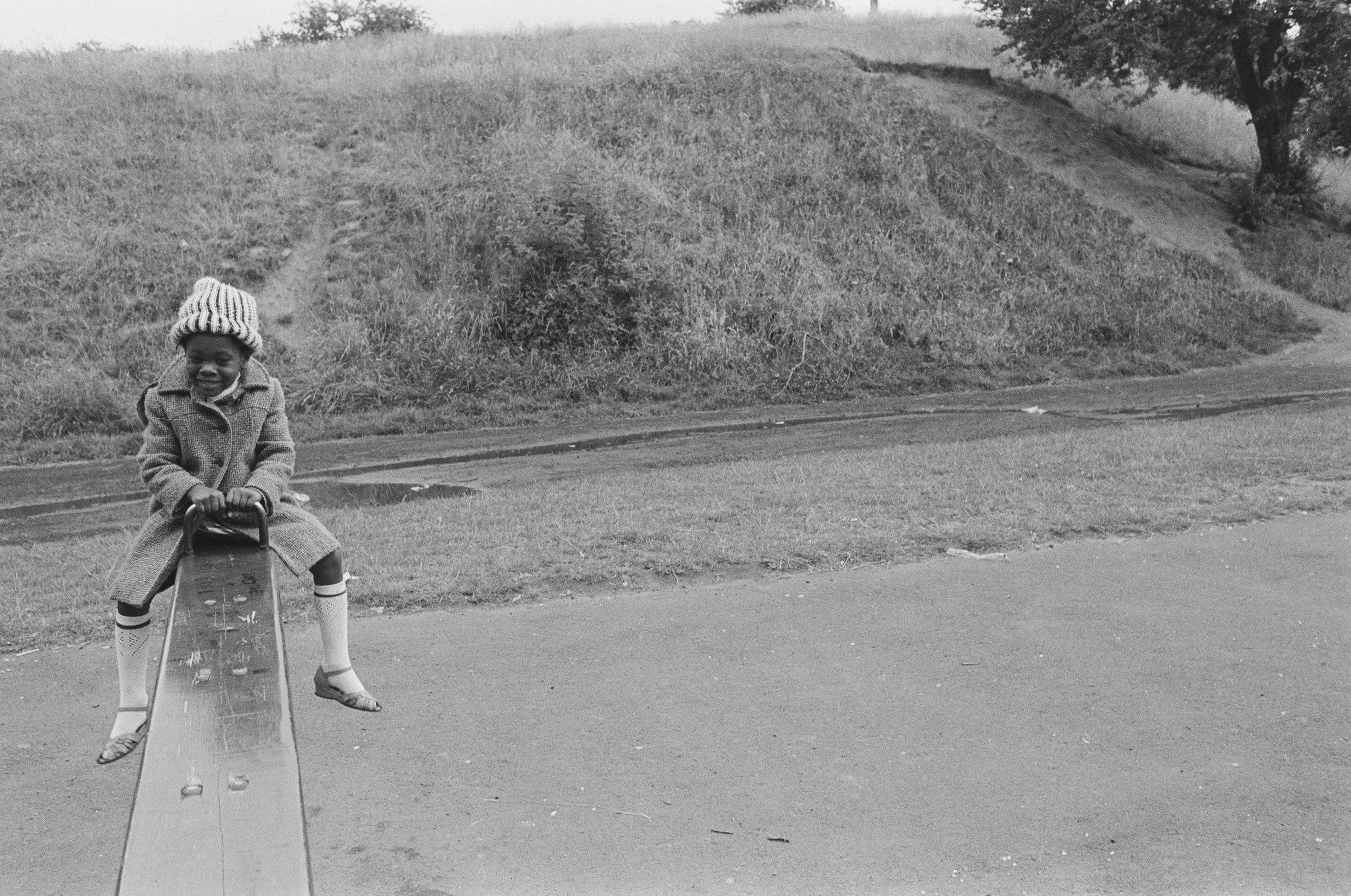 Image description: black and white landscape photograph of a dark skinned child with dark hair on a seesaw. They are wearing a light and dark striped woollen hat, a light winter coat, long white socks and open toed shoes. She is holding on to the han