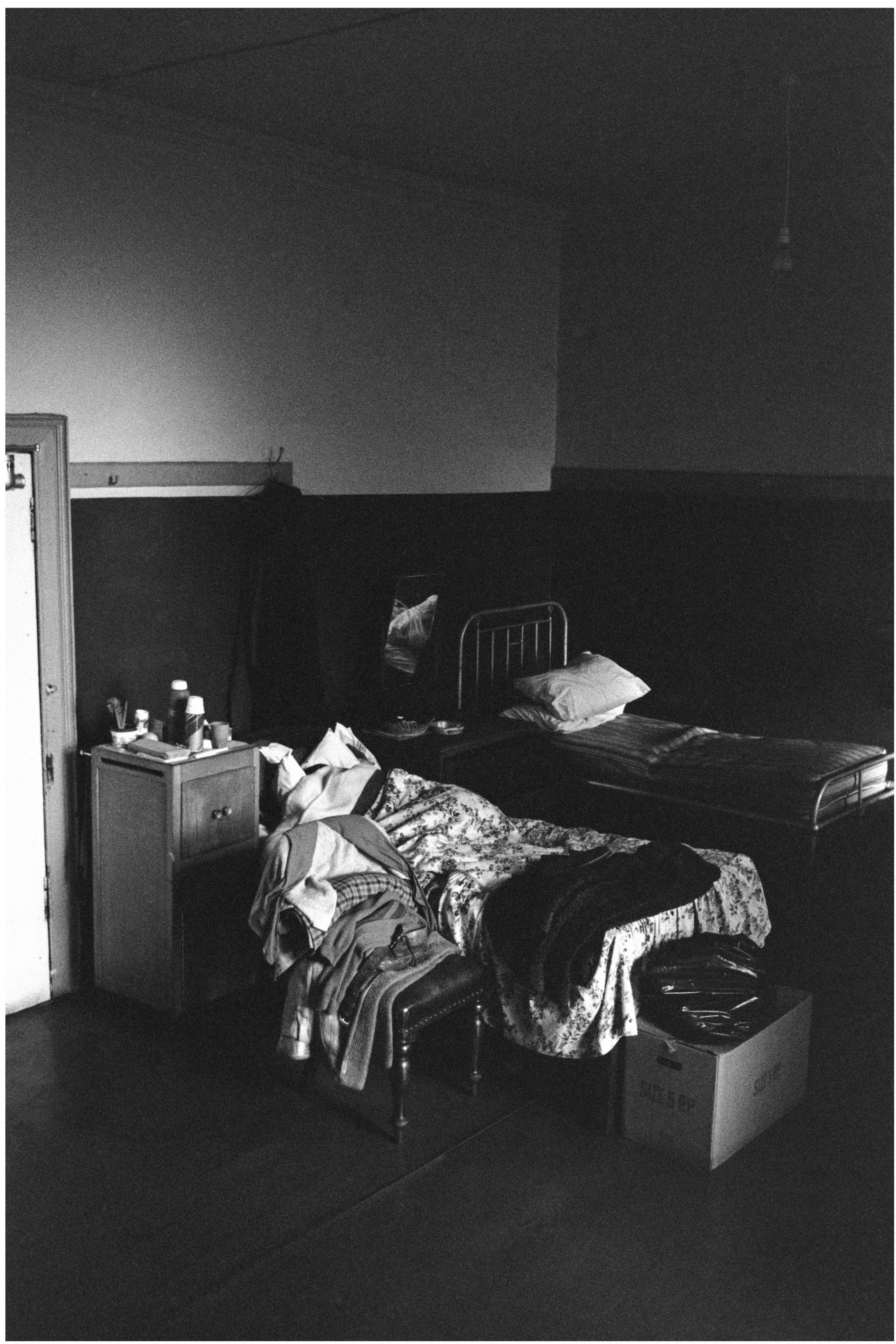 Image description: black and white portrait image of a room at the Victoria Hostel. The bed in the background is unoccupied and stripped. A person lies in the bed in the foreground. The bed has a floral duvet. There is a chair covered in clothes, and