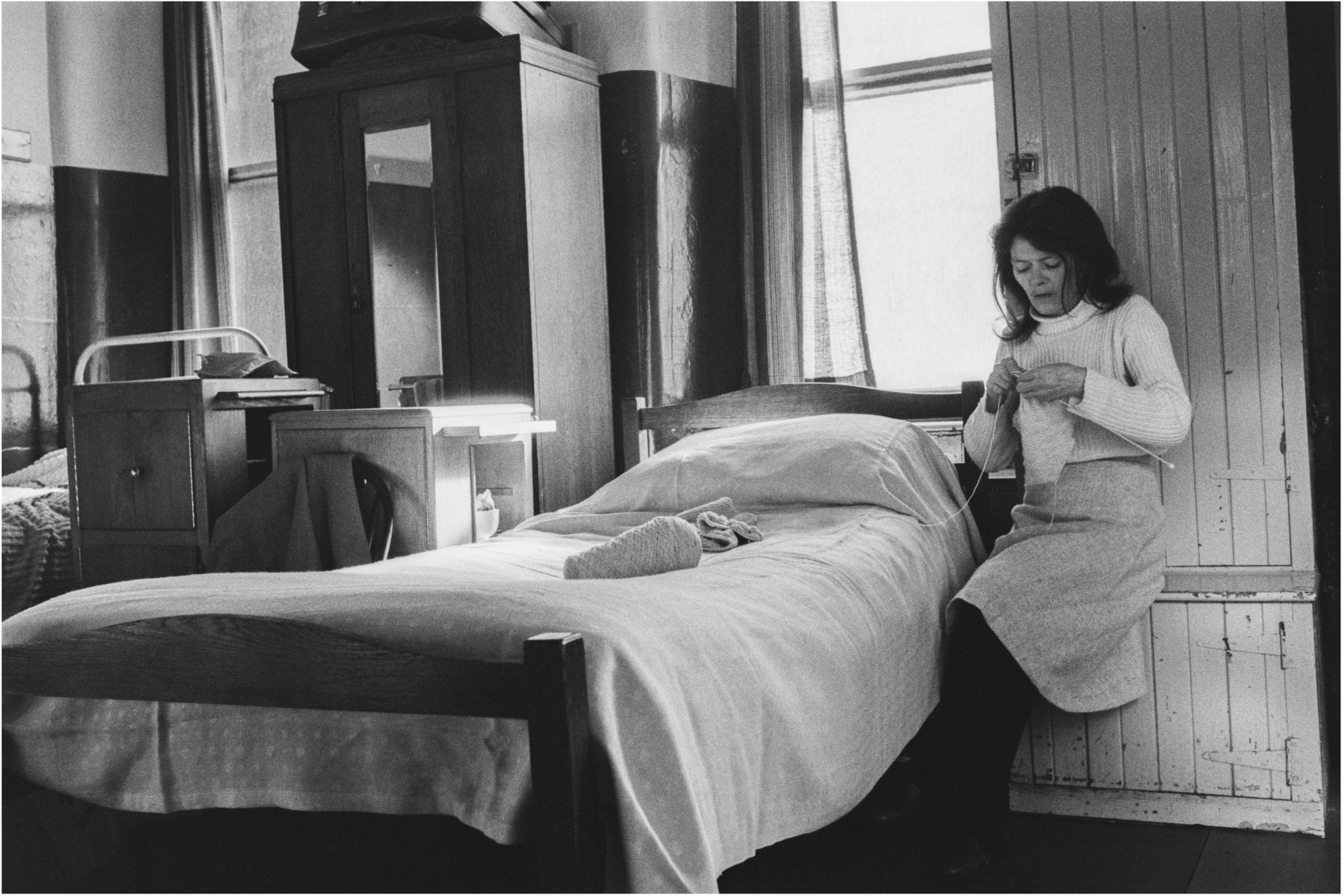 Image description: black and white landscape photograph of a room at the Victoria Hostel. A woman with light skin and long dark hair wearing a white jumper, light coloured skirt, and black tights sits on a wooden seat next to a neatly made white bed.