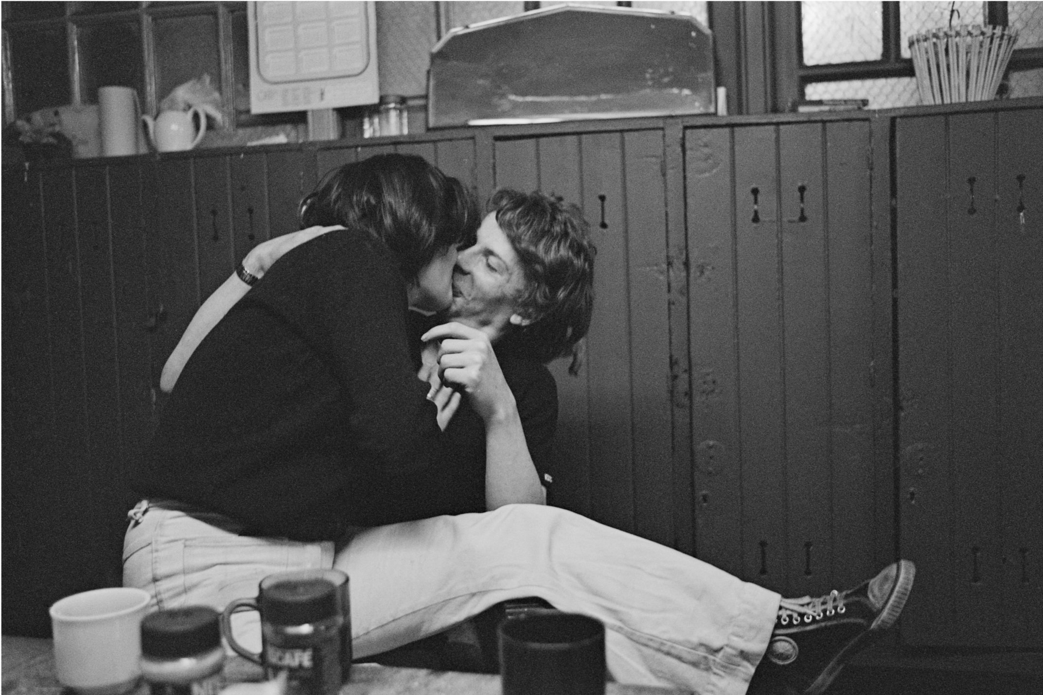 Image description: black and white landscape photograph of a couple at the Victoria Hostel. A man and a woman, both in black shirts and white trousers, kiss at a table in front of a series of floor to ceiling cupboards. The woman sits on the man's la