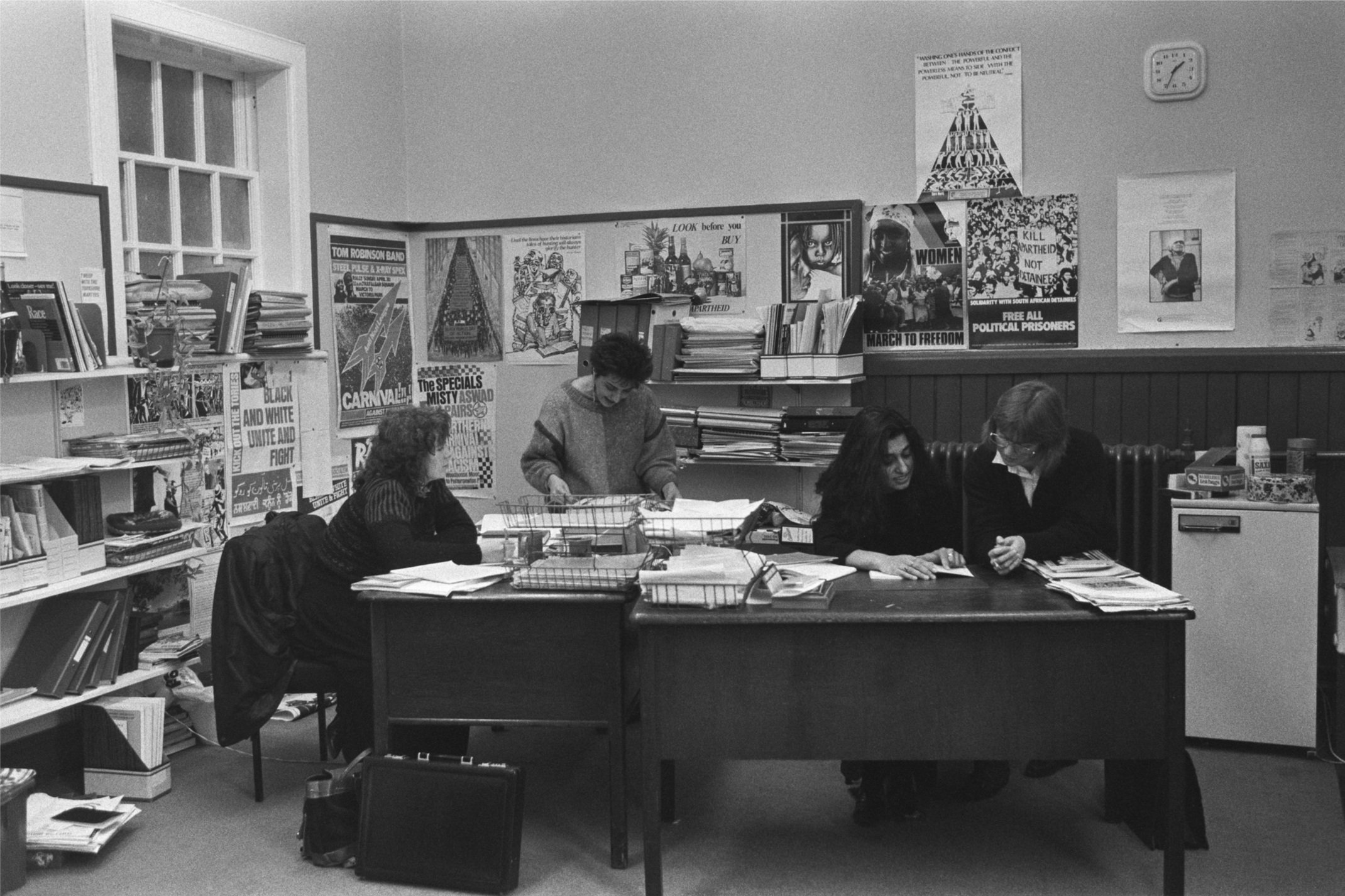 Image description: black and white landscape photograph of a group of women working in the Shakti office. Four women read documents and converse around a large brown desk. There are poitical posters covering the walls, and papers, files, and books on