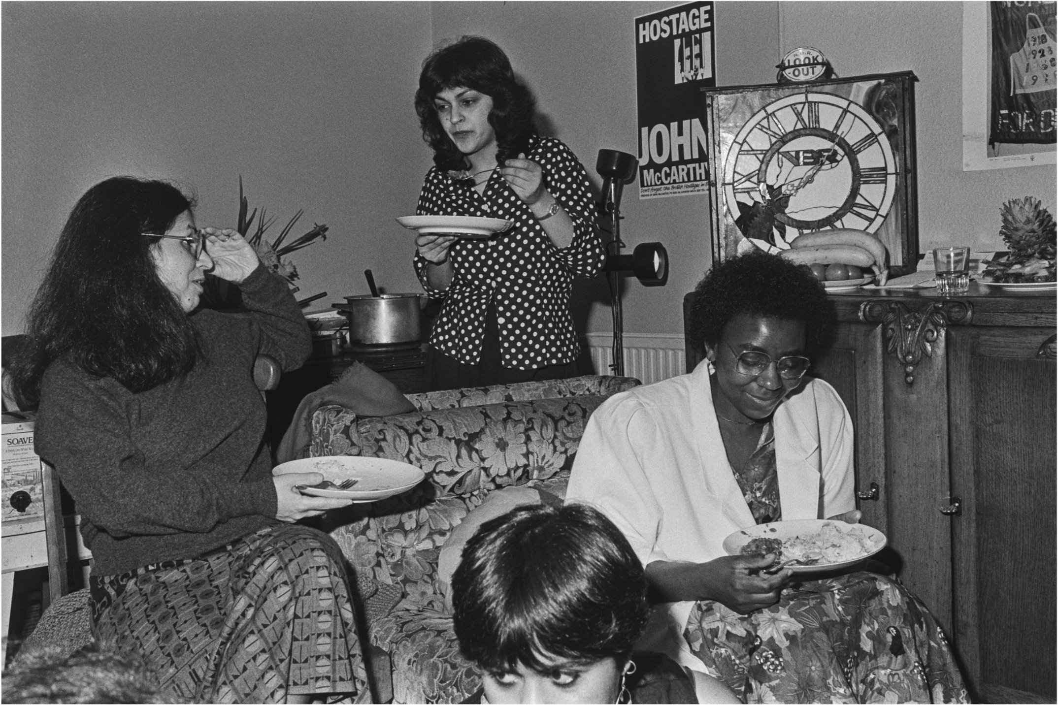 Image description: black and white landscape photograph of four women eating dinner in a living room. On the right, a woman with dark skin, short dark hair, and glasses, wearing a white blazer and patterned dress, sits on a patterned armchair and smi