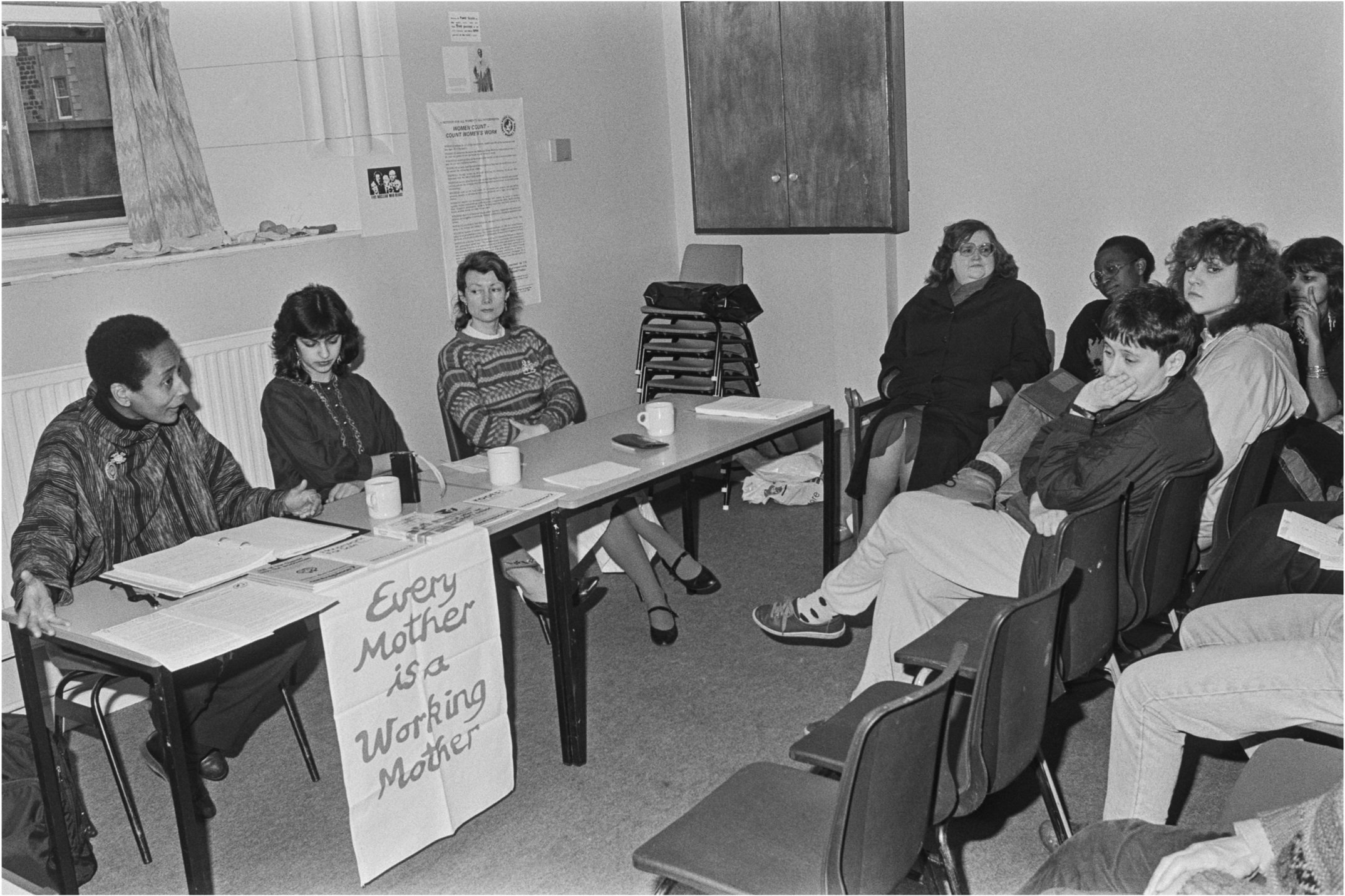 Image description: black and white landscape photograph of a Shakti meeting. Three women sit behind a table, watched by a larger group of women sitting on rows of chairs. Two of the women are listening to a third woman, who is talking to the room. Sh