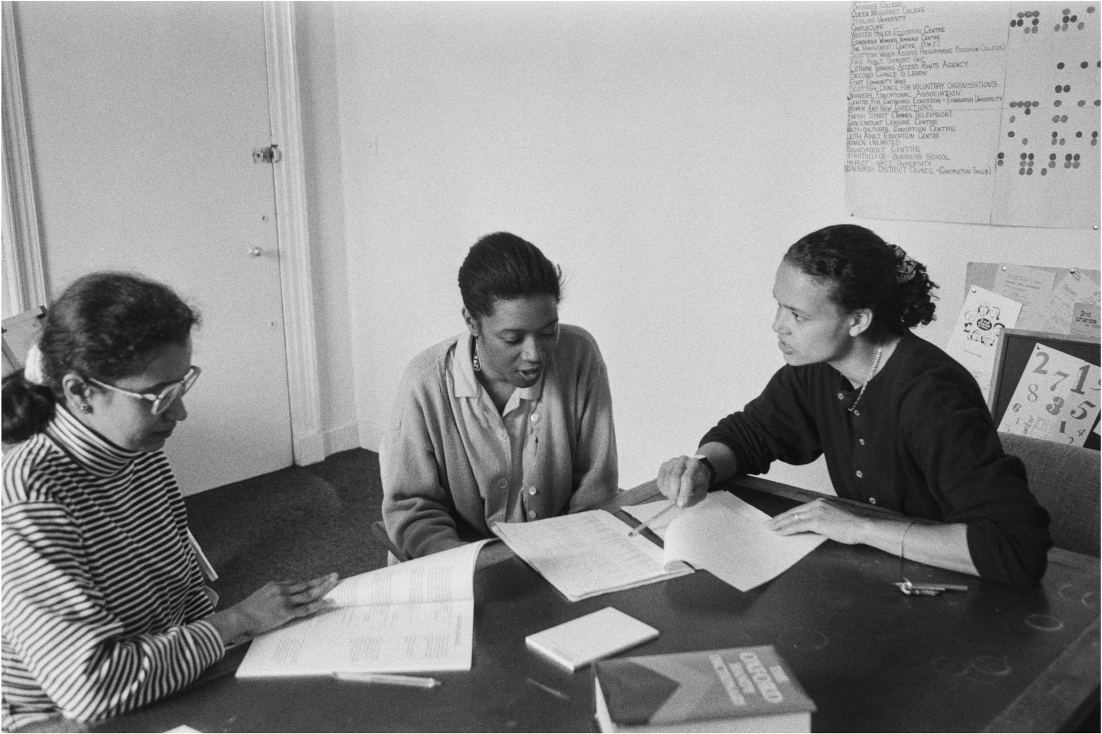 Image description: black and white landscape photograph of a Shakti meeting. Three women sit around a table. Two of the women are listening to a third woman on the right, who is talking to through a document in front of them. She has medium light ski