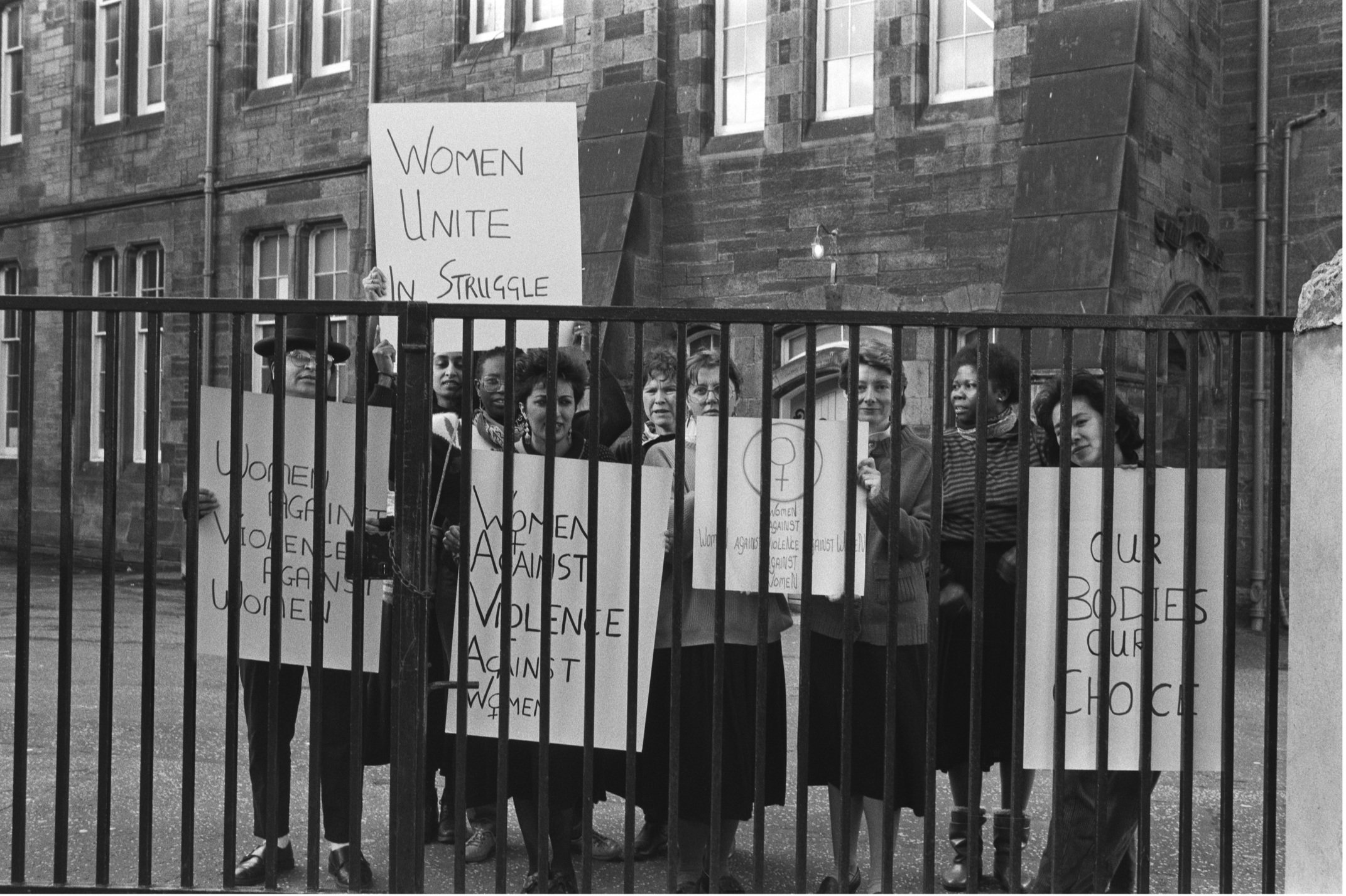 Image description: black and white landscape photograph of a group of woman behind a set of dark railings. They hold placards, which read ' WOMEN AGAINST VIOLENCE AGAINST WOMEN', 'WOMEN UNITE IN THE STRUGGLE', and 'OUR BODIES OUR CHOICE'. They are sm