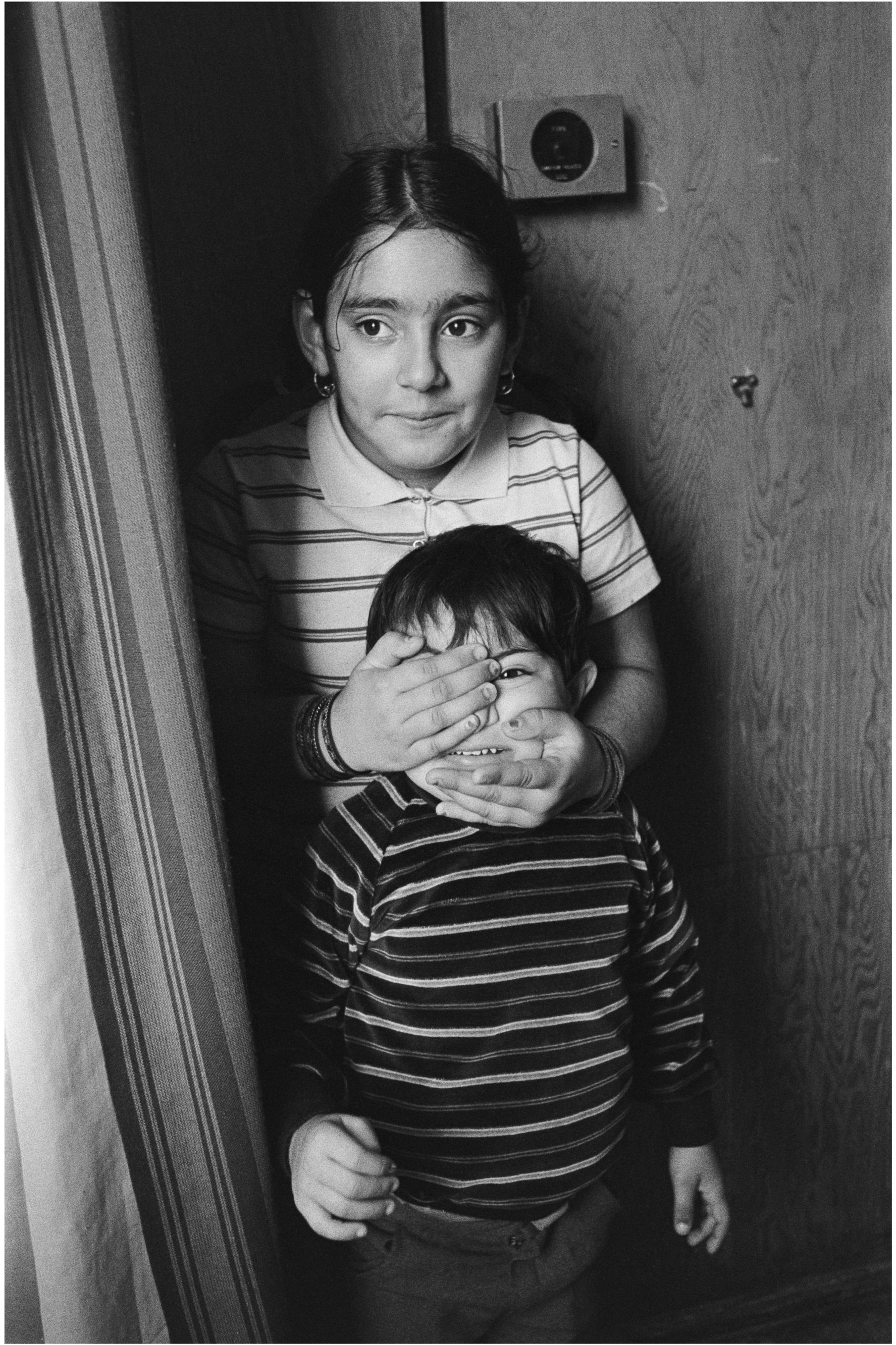 Image description: Black and white protrait of two children standing beside a striped curtain in front of a wooden wall. The taller child has medium light skin and dark hair, and is wearing a striped light and dark collared polo shirt. There are brac