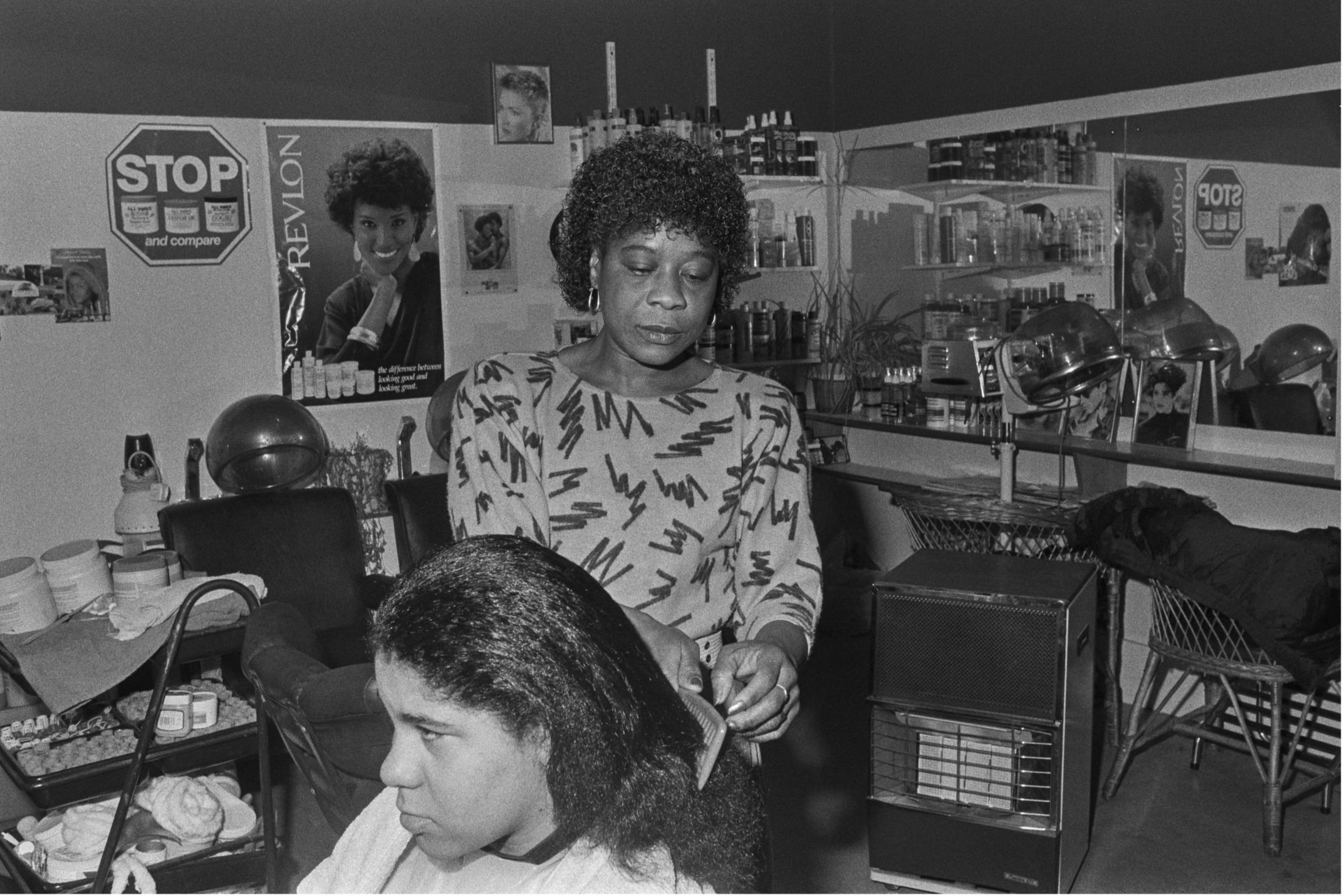 Image description: black and white landscape photograph a a hairdressing salon. A woman with dark skin and short dark hair wearing a grey jumper patterned with jagged lines combs the hair of a second woman sitting in a barbers chair next to a set of