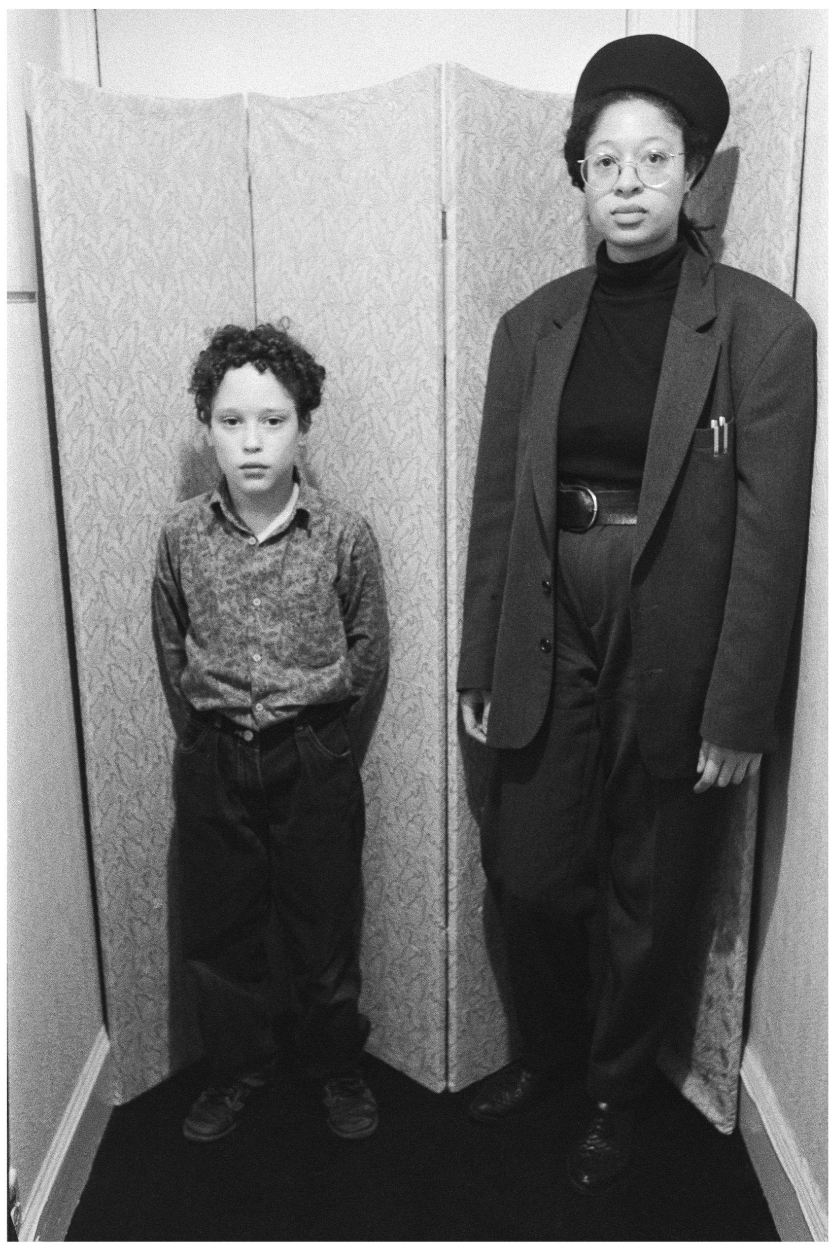 Image description: black and white portrait photograph of Sandra George and her son Tyler. They are standing in front of a dressing screen. Tyler has light skin, short dark curly hair, and is wearing a patterned shirt and dark trousers. Sandra has me