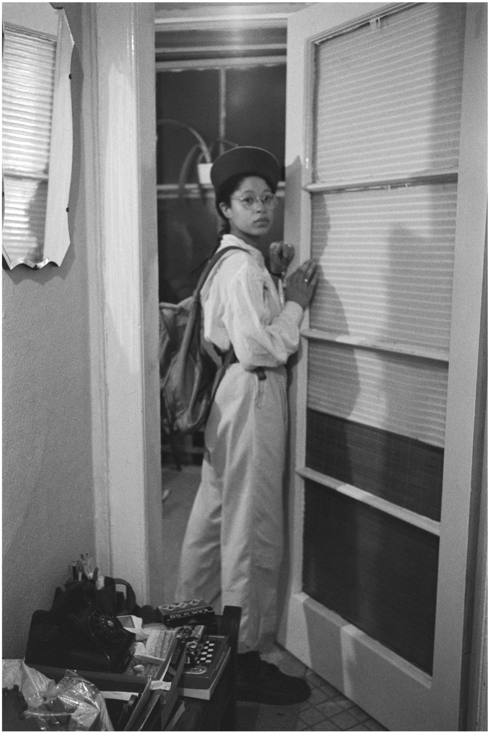 Image description: black and white portrait photograph of Sandra George. She is standing in a doorway of her flat. Sandra has medium light skin, long dark hair in a single braid, and is wearing a white shirt, light coloured trousers, a dark pillbox h