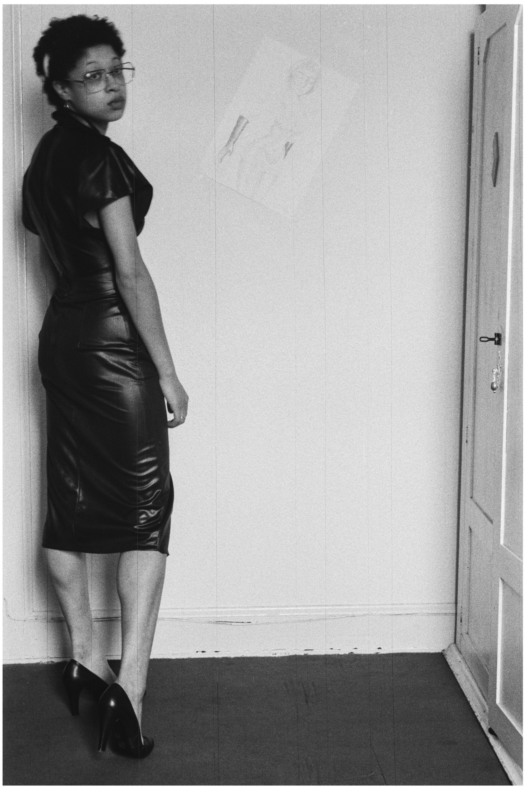 Image description: black and white portrait photograph of Sandra George at home. Sandra George has medium light skin and short dark hair, and is wearing a dark fitted dress, dark high heels, and glasses. She is facing away from the camera, but is loo