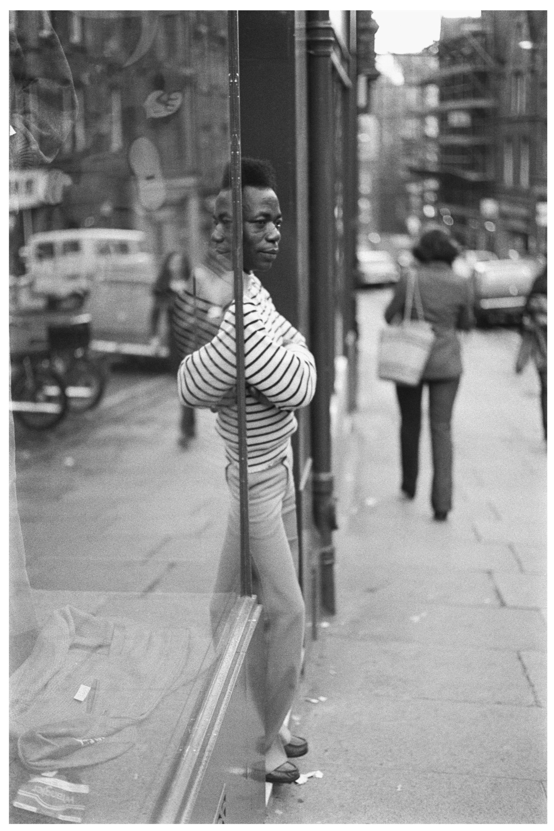 Image description: A black and white portrait of a man in profile leaning against a shop doorway looking out into the street. The man has dark brown skin and a short dark flat top hair cut, and is wearing a striped white and black jumper and light co