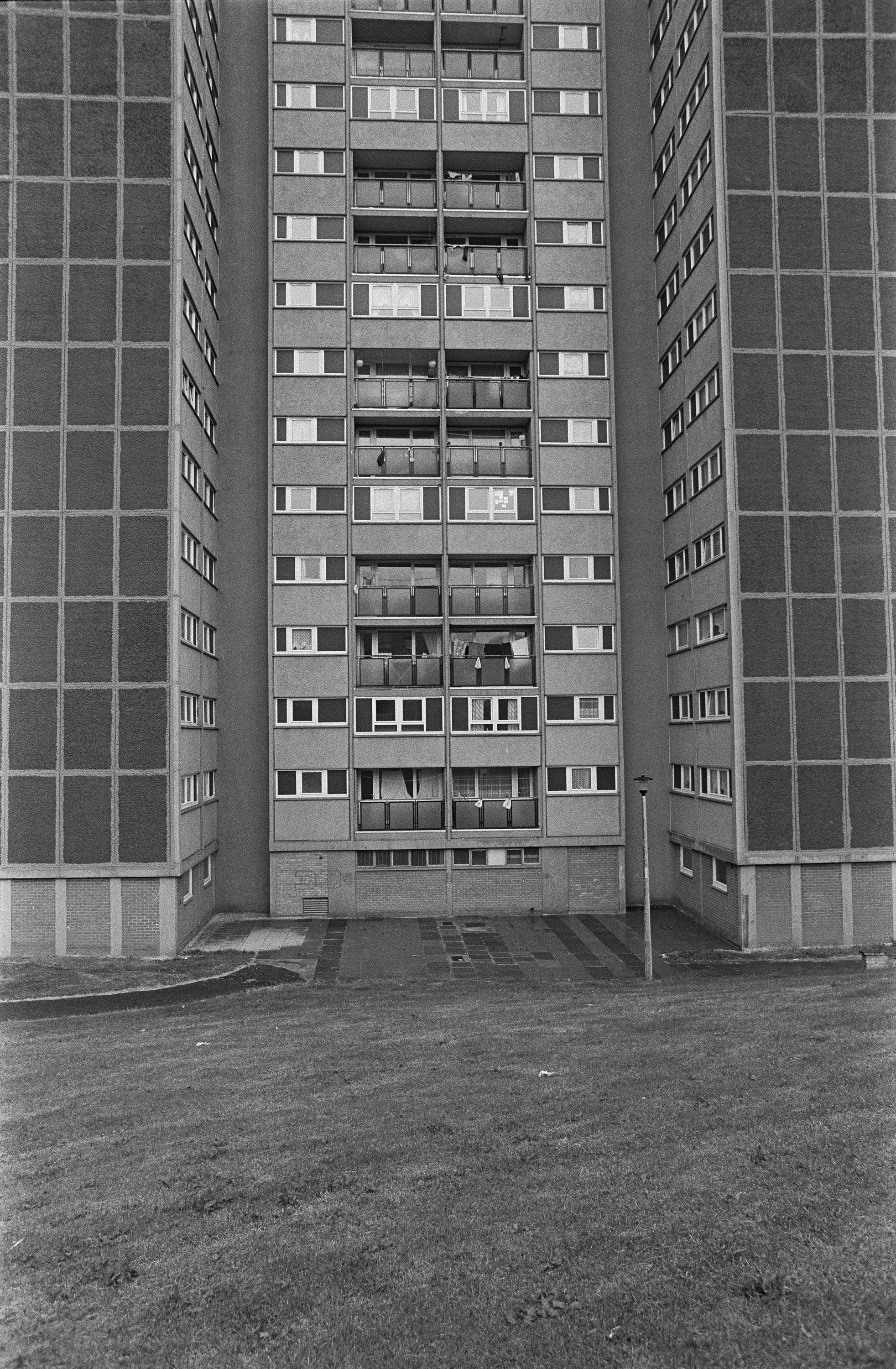 Image description: black and white portrait image of high rise flats in Oxgangs. The building has dark patterned sections on the front elevation, and many small windows and balconies rising up out of sight.