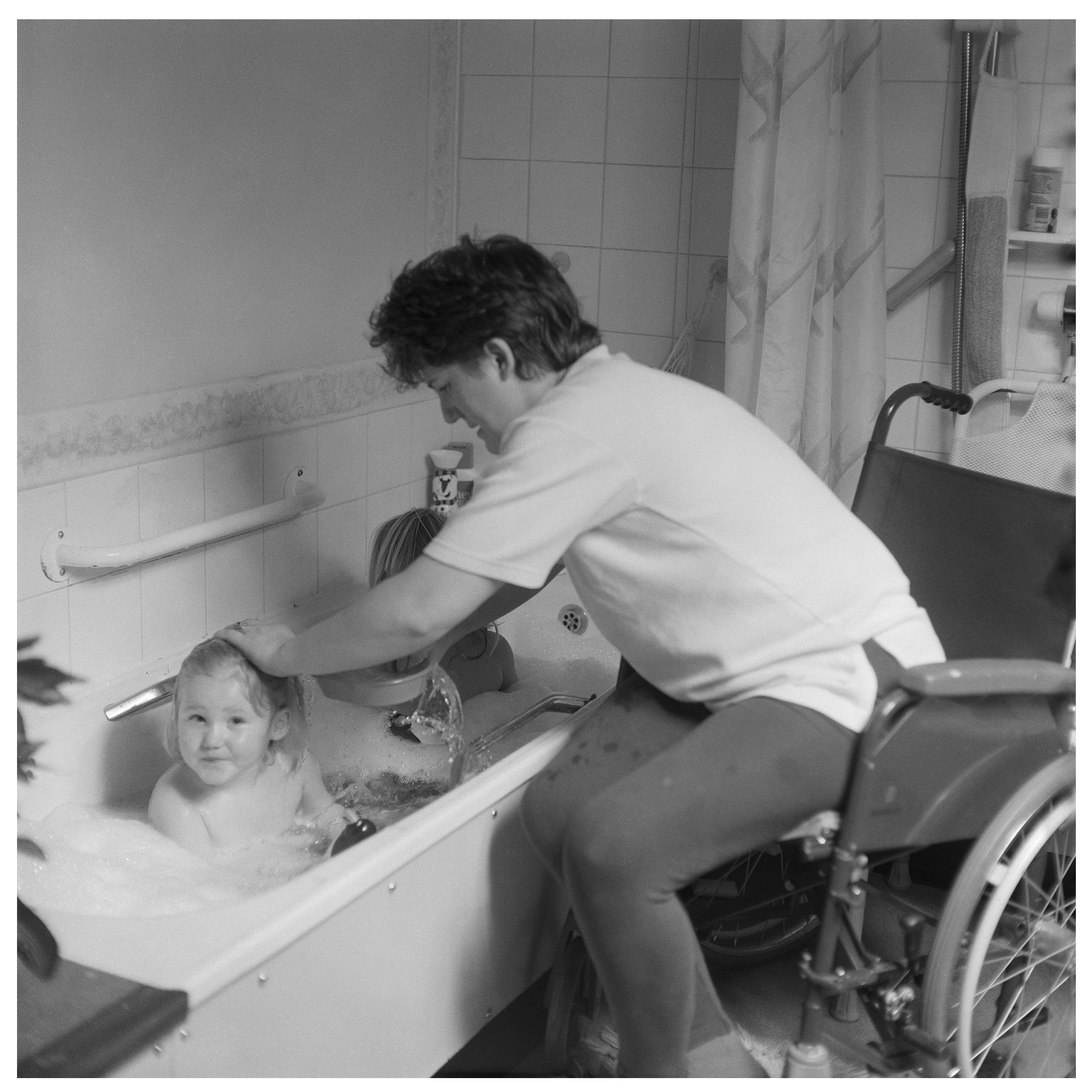 Image description: black and white image of a woman using a wheelchair and bathing two children in a bath tub. The woman on the right of the image has light skin and short dark hair, and is wearing a white T shirt and grey trousers. She sits in a dar