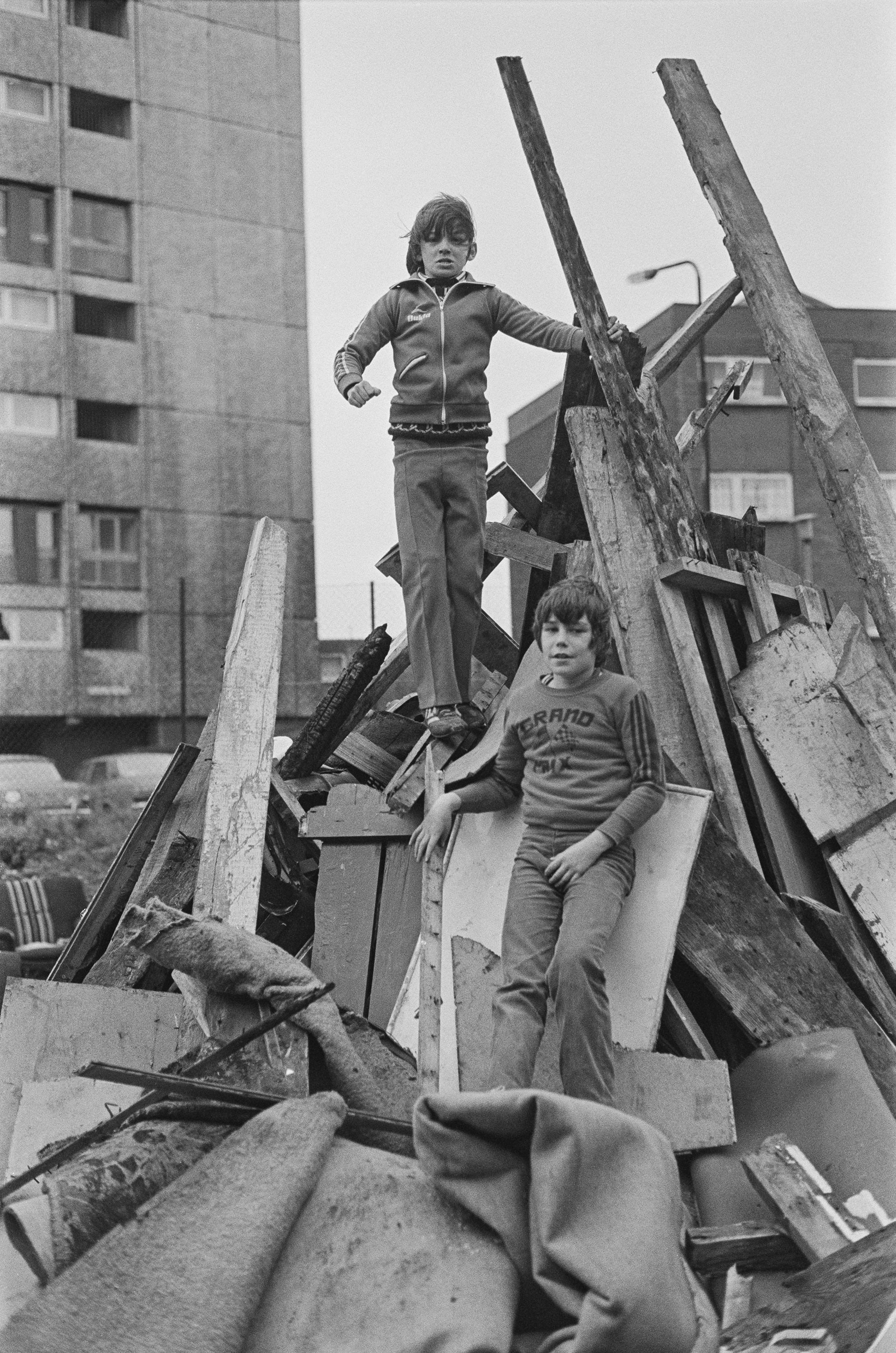 Image description: black and white portrait photograph of two children standing amidst a pile of wood for a bonfire. One has light skin, mid length dark hair, and wears a grey sweatshirt and jeans, and is leaning against pieces in the middle of the p