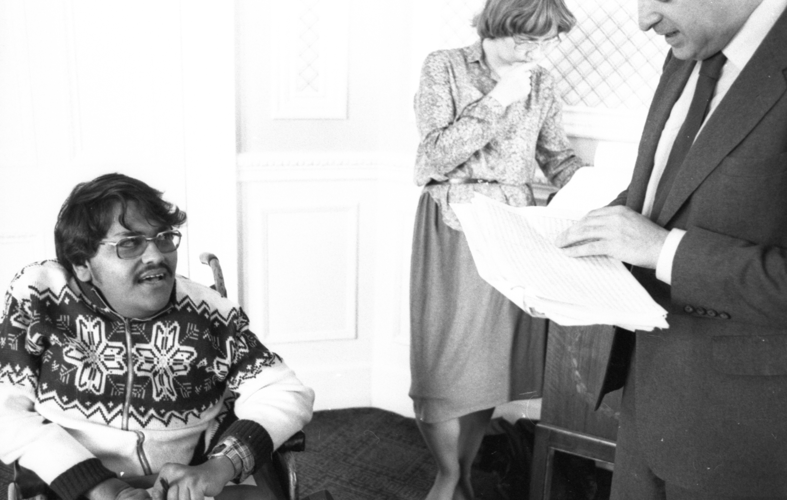 Image description: black and white landscape image of three people. On the left, a man with medium light skin, dark hair, a moustache, and glasses wears a patterned knitted zip up jumper and a wristwatch. He is using a wheelchair. He is speaking to a