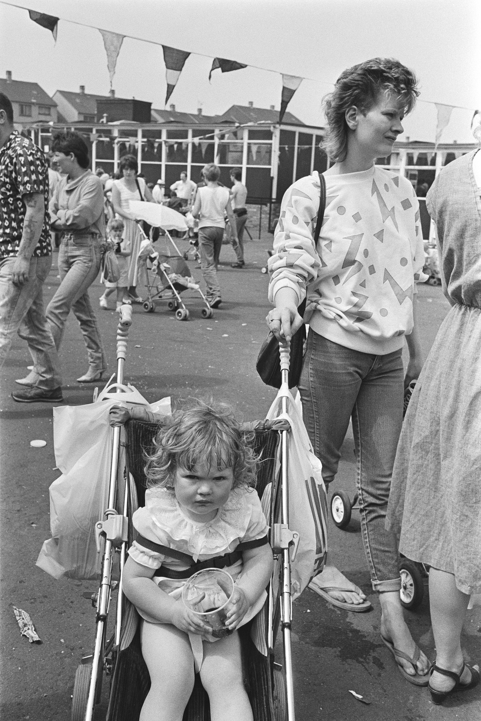 Image description: black and white portrait of a child in a buggy. The child has light skin and light mid length hair, and is wearing a frilly white top. She is strapped into the buggy with dark straps, and there are plastic bags slung over both hand
