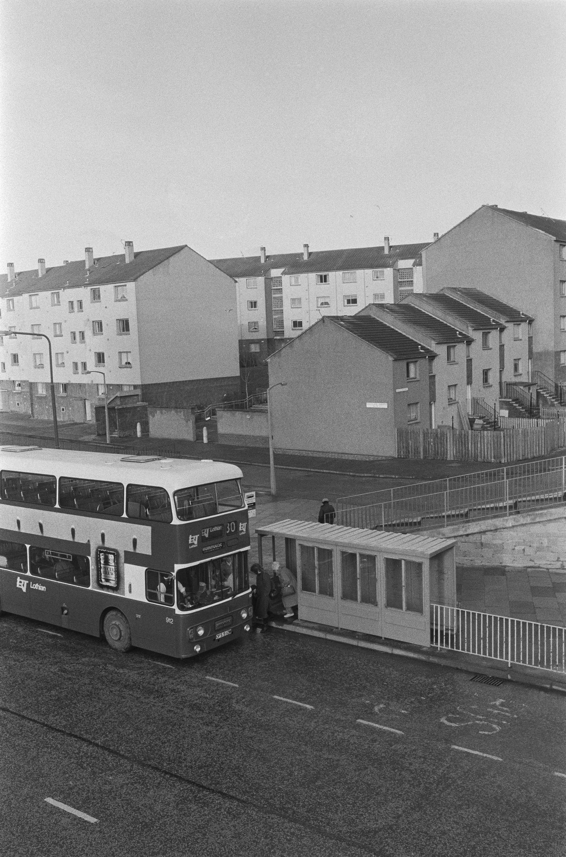 Image description: black and white portrait image of a bus stop in Wester Hailes from an elevated position. The number 30 bus is parked at the stop, and two women are boarding. The bus stop is at an intersection.