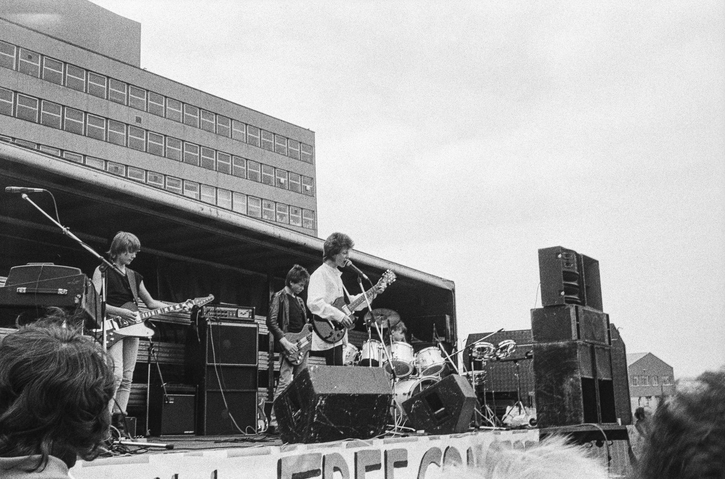 Image description: black and white image of a band peforming onstage. The stage is raised, with speaker towers on either side and a dark backdrop with silver metallic stripes. A banner hangs at the front. On the left of the image, a man with light sk