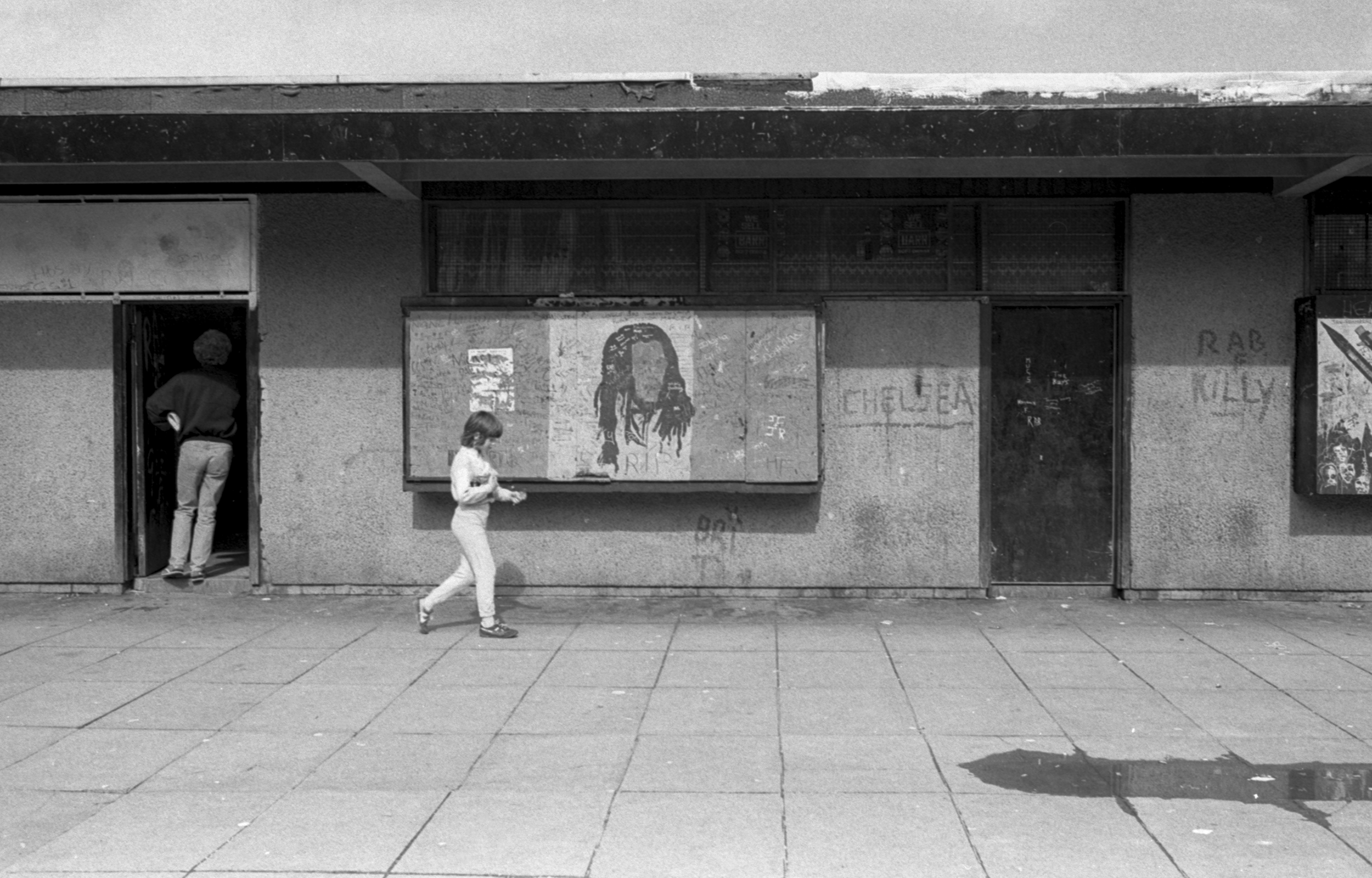 Image Description: A black and white landscape photograph showing the shopfronts along Niddrie Mains Road. In the centre of the frame is boarded up front with a artwork of a man with dreadlocks. A child walks past the painting holding something unde